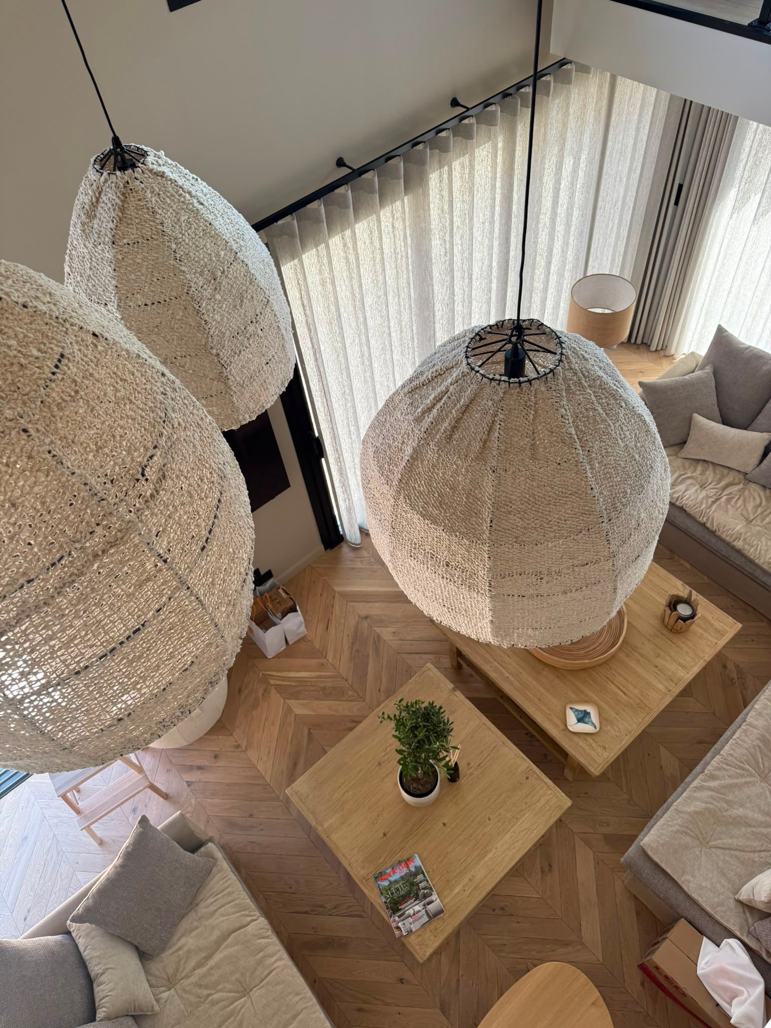 Salon moderne avec des suspensions en osier, des tables en bois, des canapés gris et de grandes fenêtres ornées de voilages