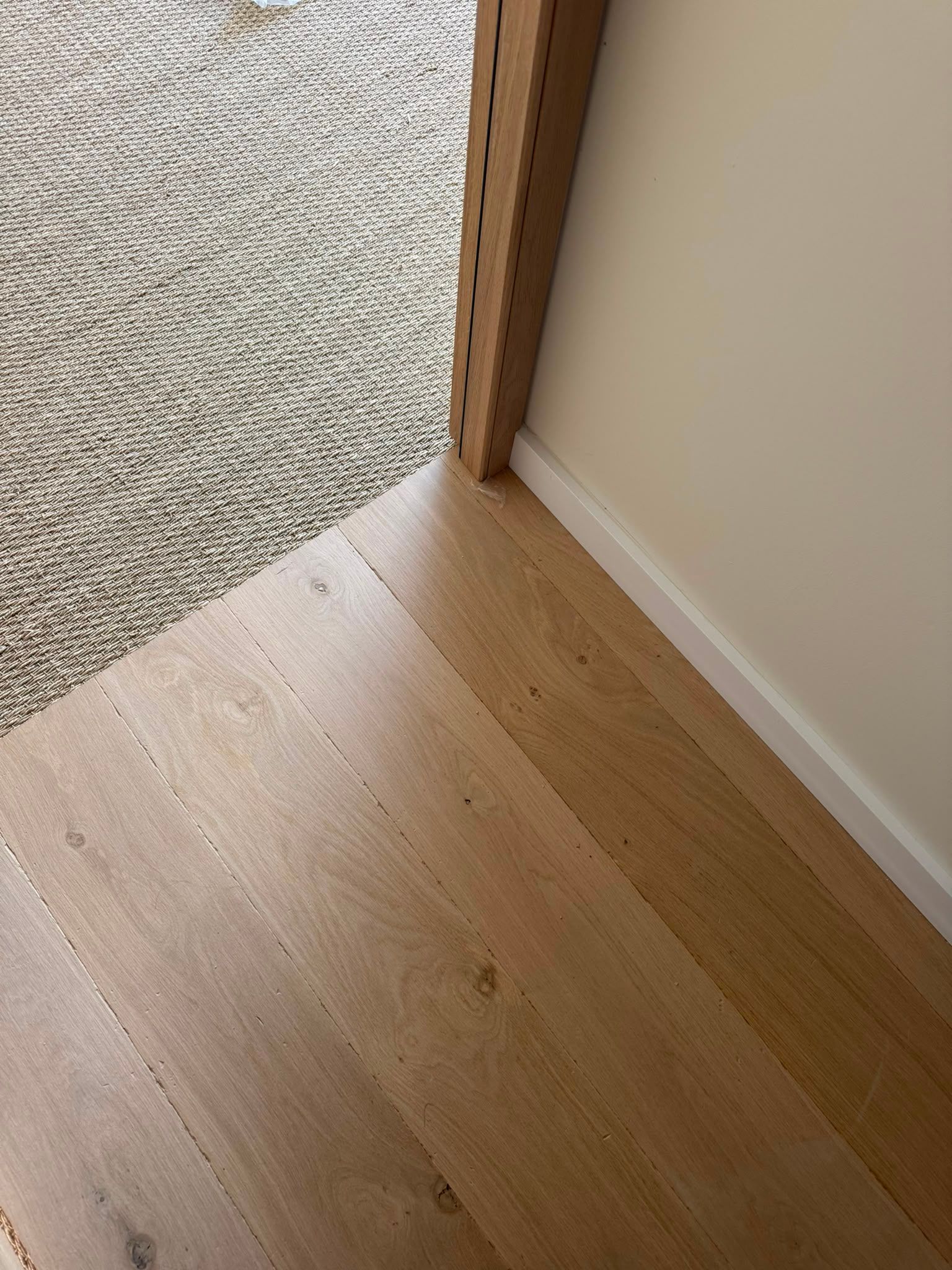 Coin d’une pièce avec un parquet clair, un tapis beige et un poteau en bois contre un mur blanc.