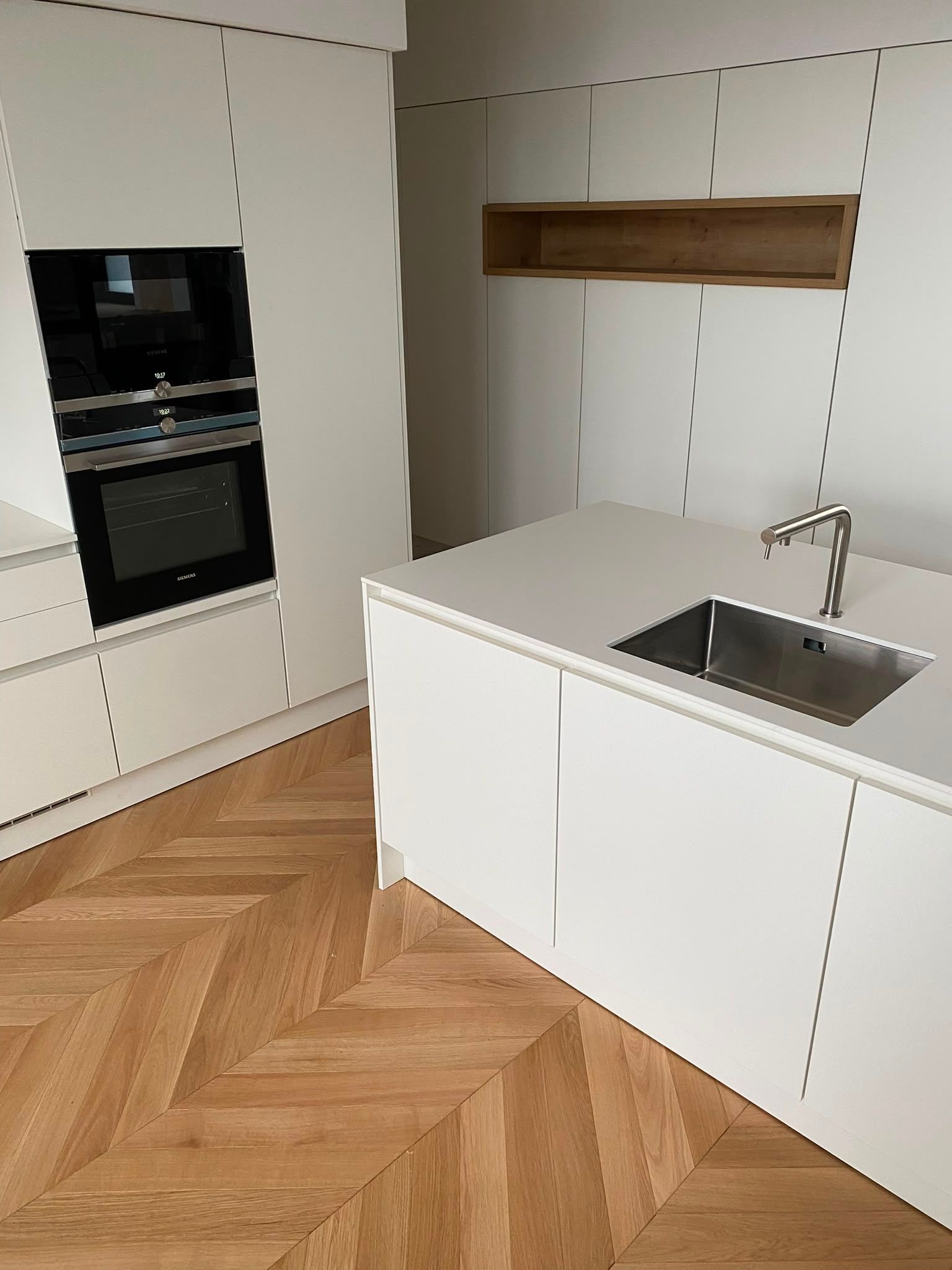Cuisine moderne blanche avec four encastré, évier dans l’îlot et parquet à chevrons.