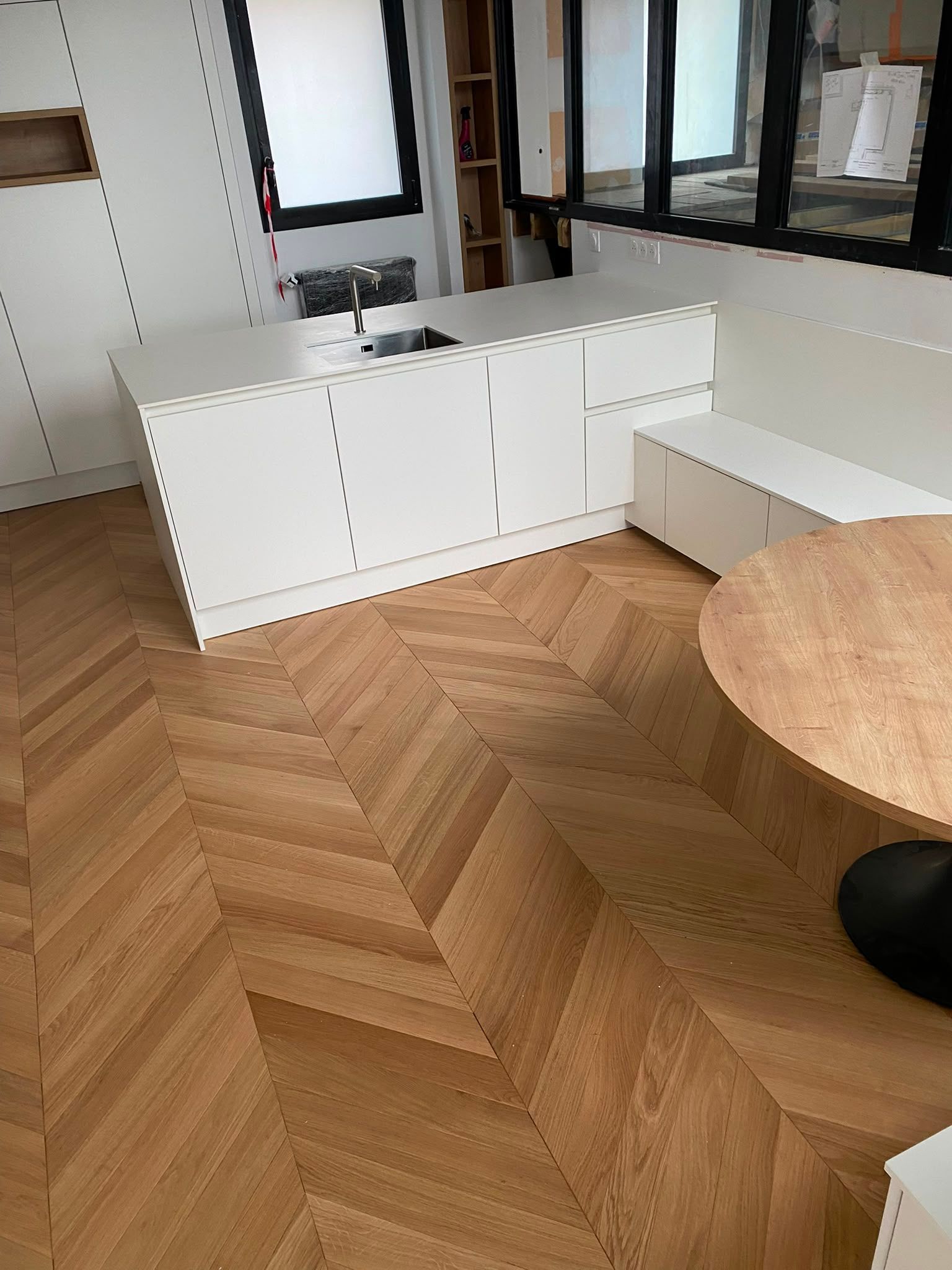 Pièce lumineuse avec parquet en chevrons, placards blancs et coin de table ronde en bois.