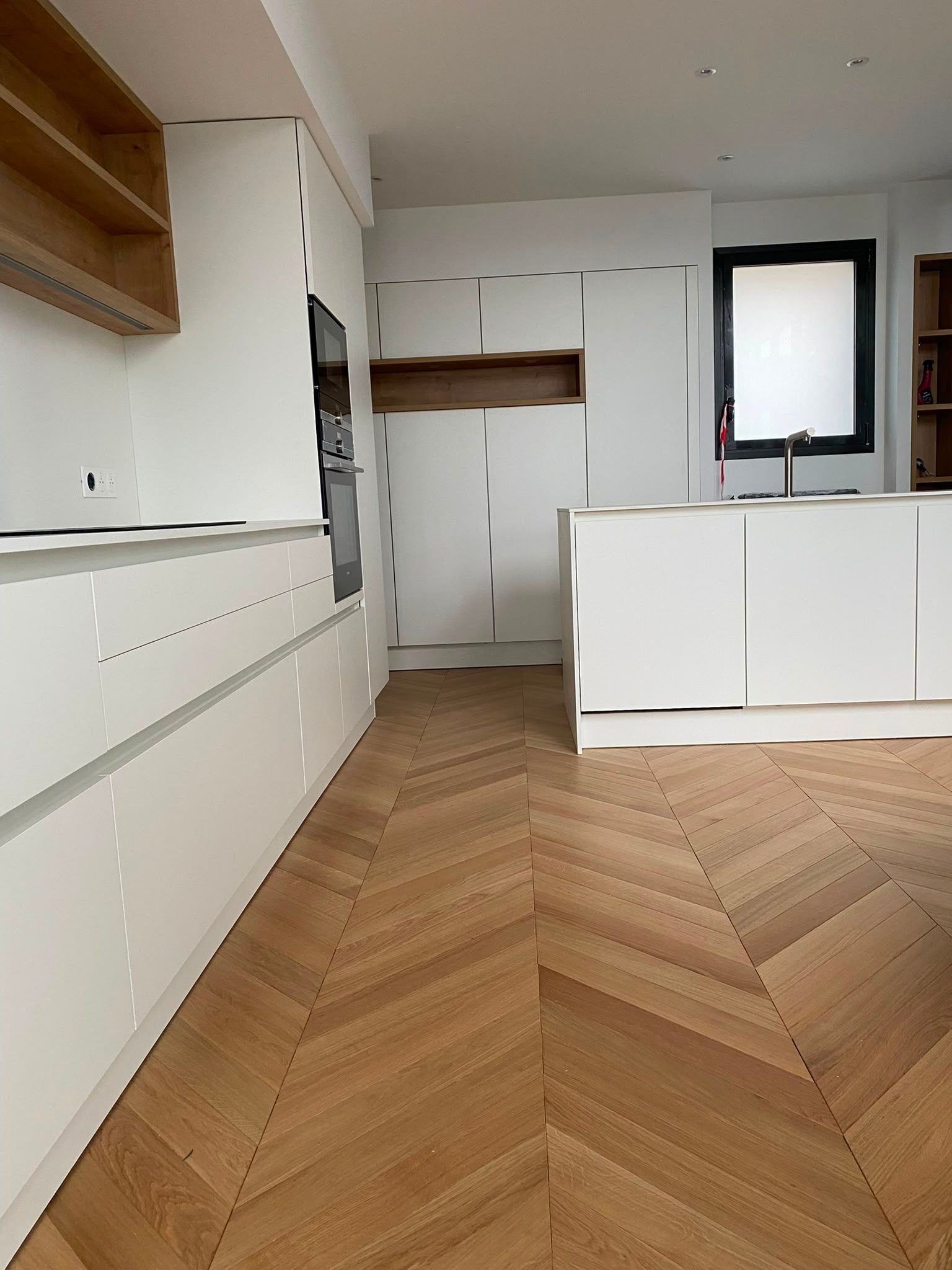 Cuisine moderne blanche avec parquet en bois à chevrons, placards intégrés, îlot central et petite fenêtre.