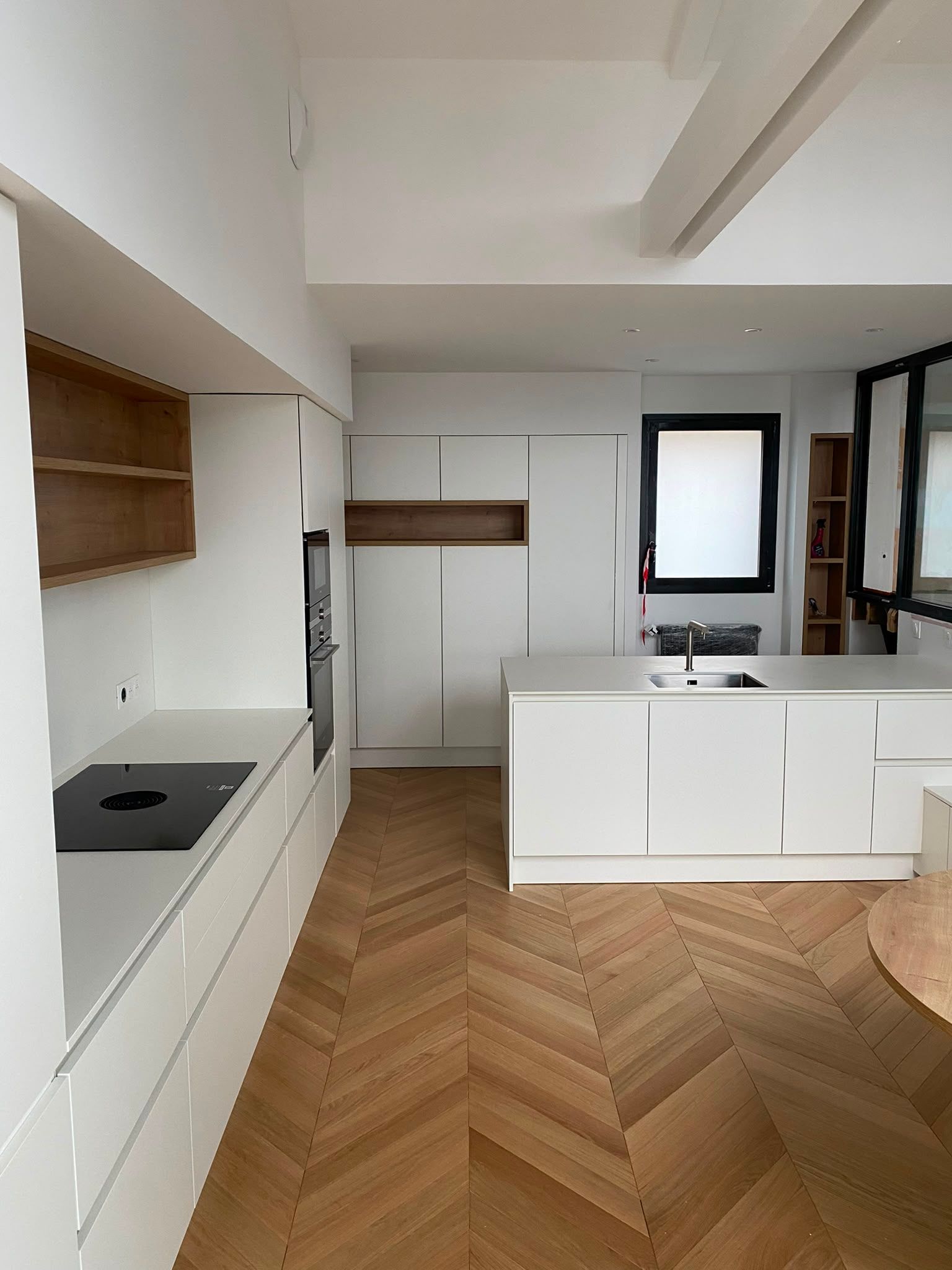 Cuisine moderne blanche avec parquet en bois à chevrons, îlot central, étagères intégrées et grande fenêtre