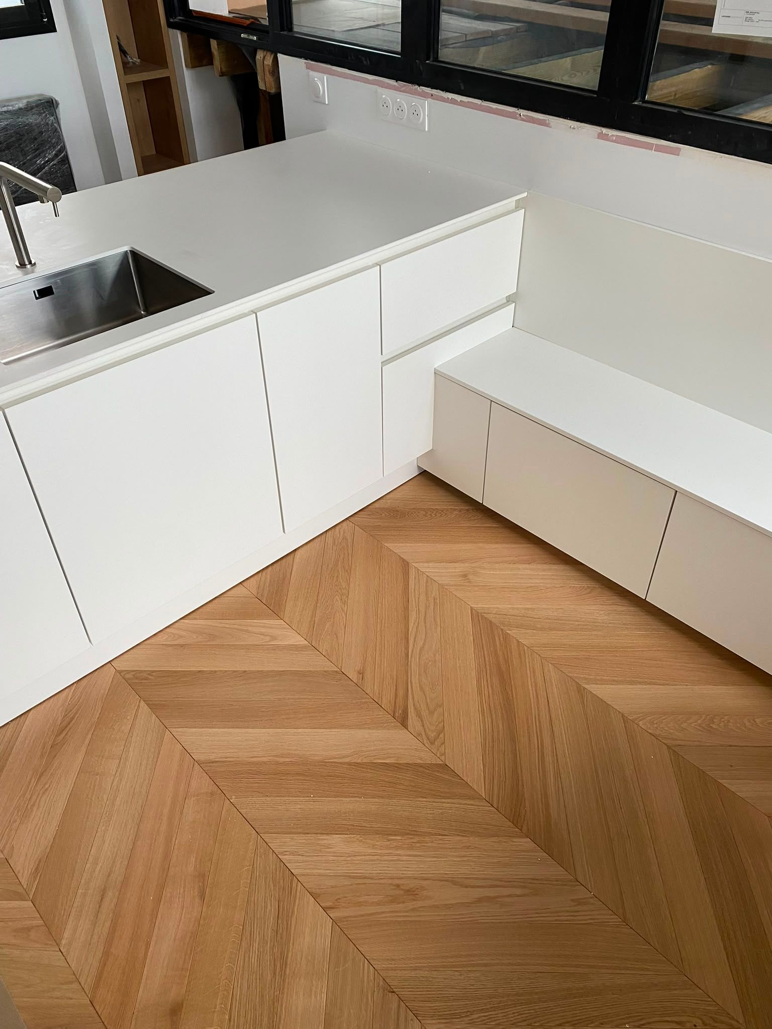 Cuisine blanche avec meubles intégrés, banquette et parquet en chevrons près d’un évier