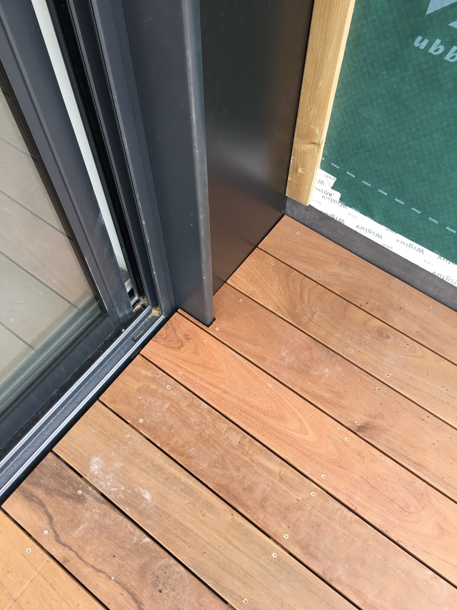 Plancher de balcon en bois, à côté d’une porte-fenêtre coulissante et d’un bardage extérieur sombre.