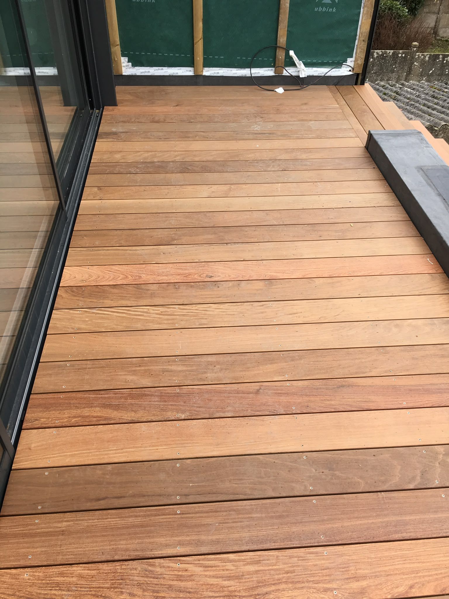 Des lames de terrasse en bois longent une rambarde en métal noir et des portes vitrées, avec un tapis gris à droite.