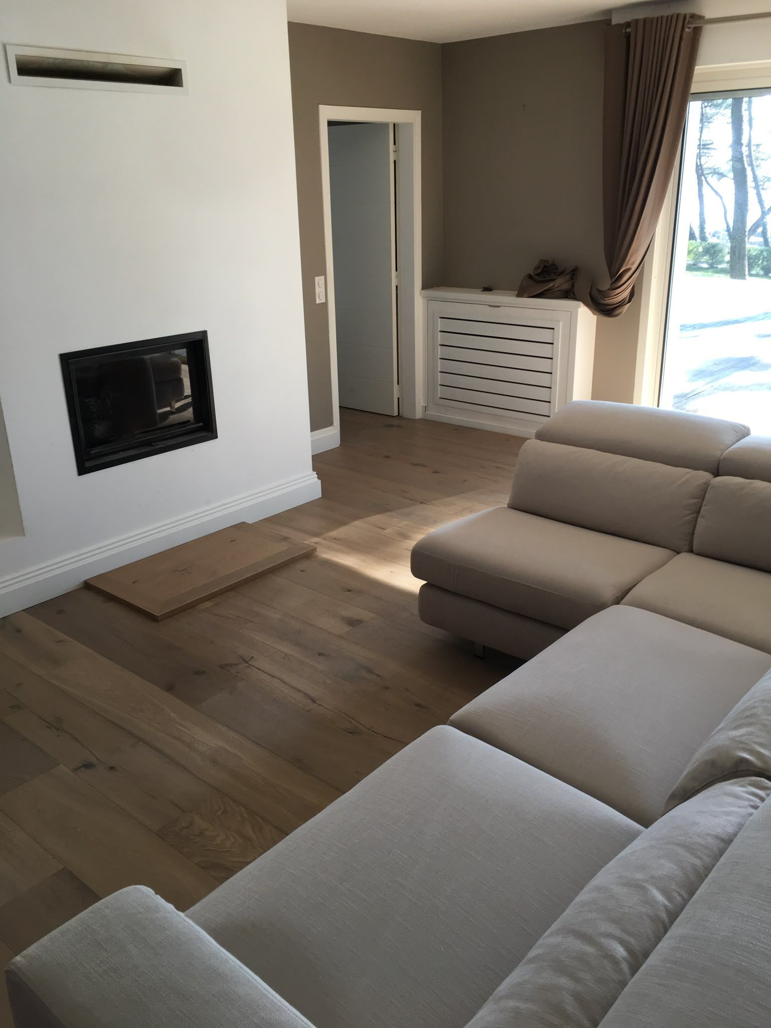Salon lumineux avec canapé d’angle beige, cheminée blanche, parquet et soleil près de la fenêtre.
