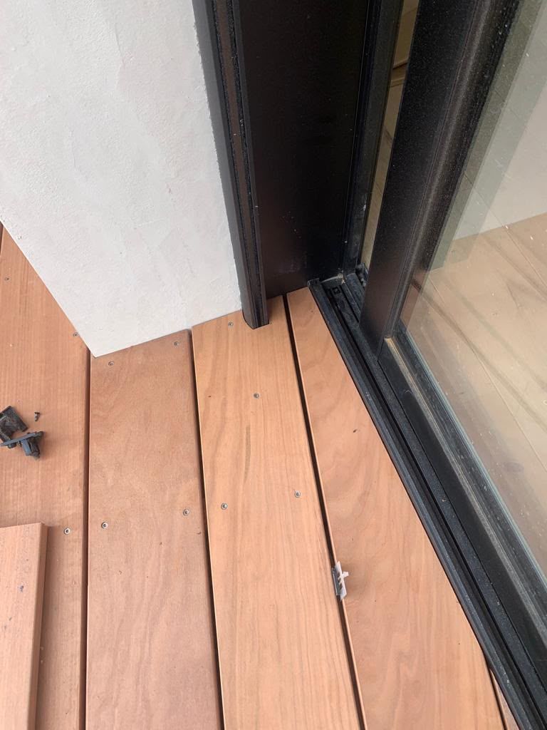 Plancher de balcon en bois près d’une baie vitrée coulissante noire et d’un mur blanc, avec un petit espace entre les lames.
