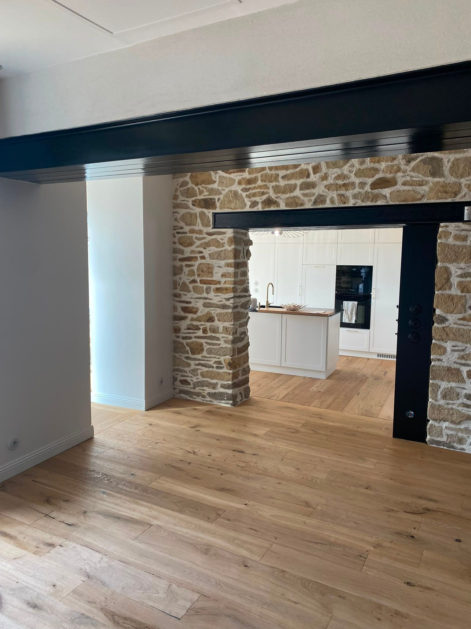 Pièce vide avec un mur en briques apparentes, des poutres noires et un parquet clair donnant sur une cuisine blanche