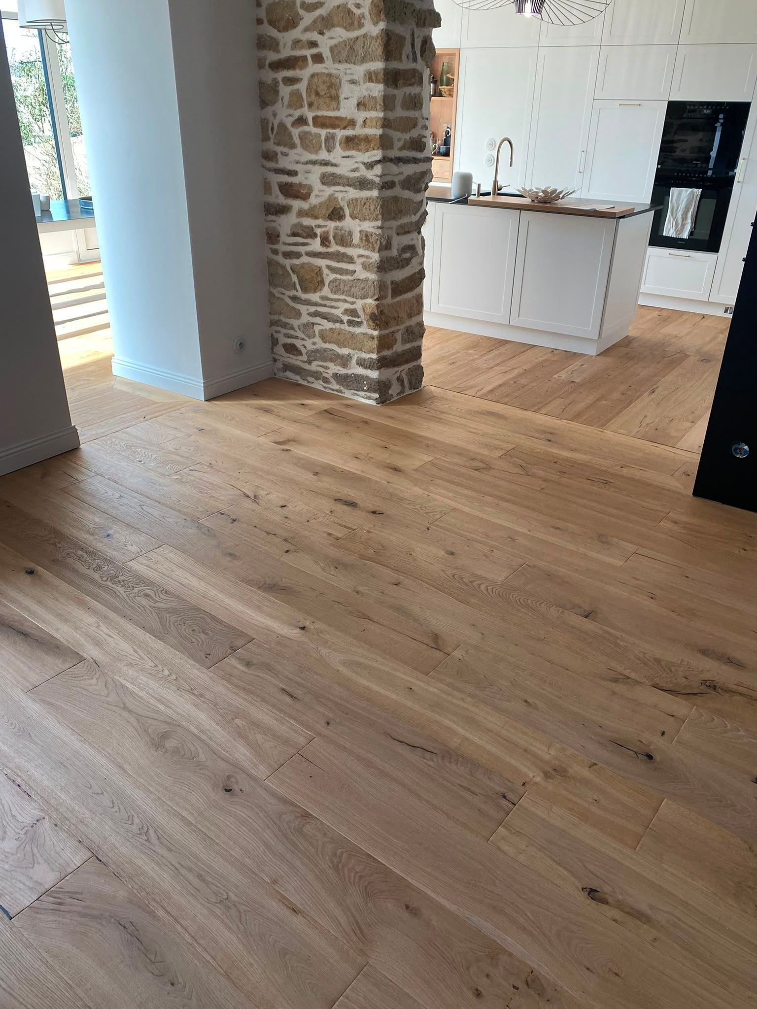 Pièce spacieuse avec un parquet clair, une colonne en pierre apparente et une cuisine blanche lumineuse en arrière-plan