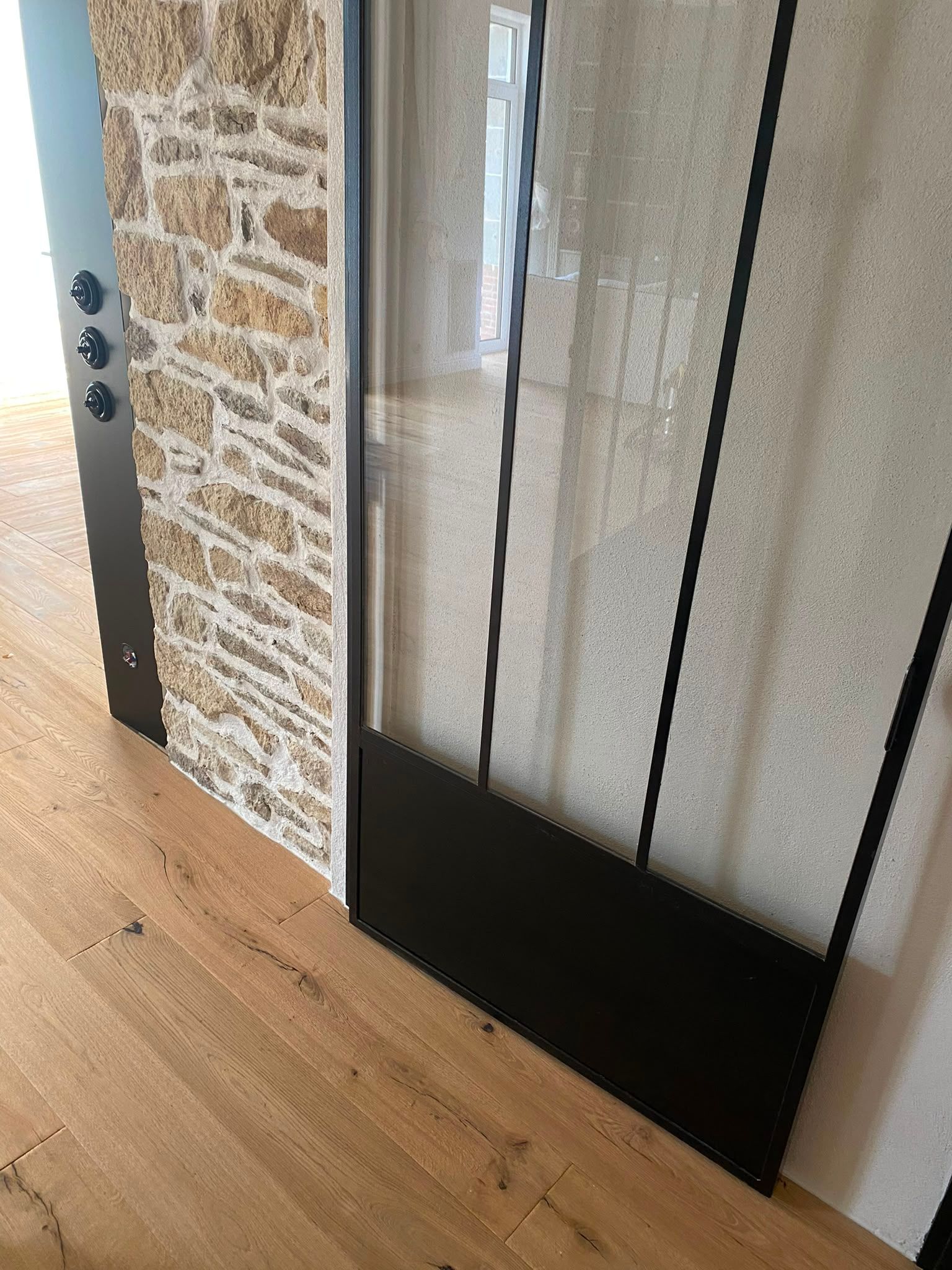 Gros plan sur une porte vitrée à cadre métallique noir, à côté d’un mur en pierre et d’un plancher en bois