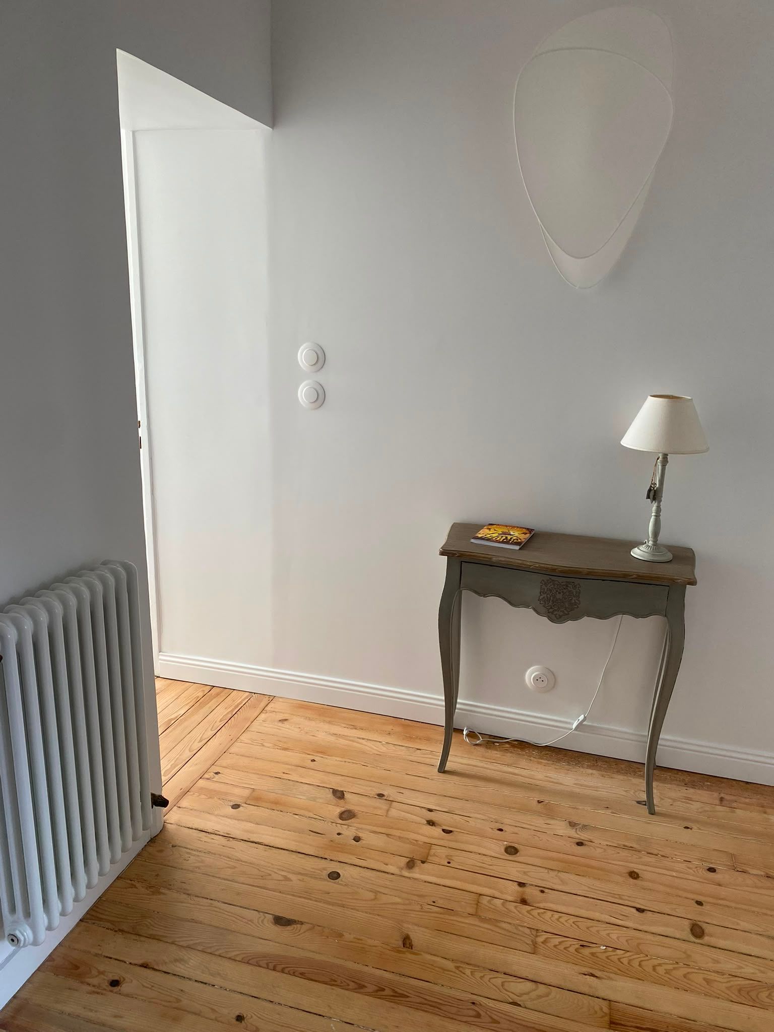 Petite pièce avec un parquet clair, des murs blancs, un radiateur, une table d’appoint, une lampe et un miroir mural.