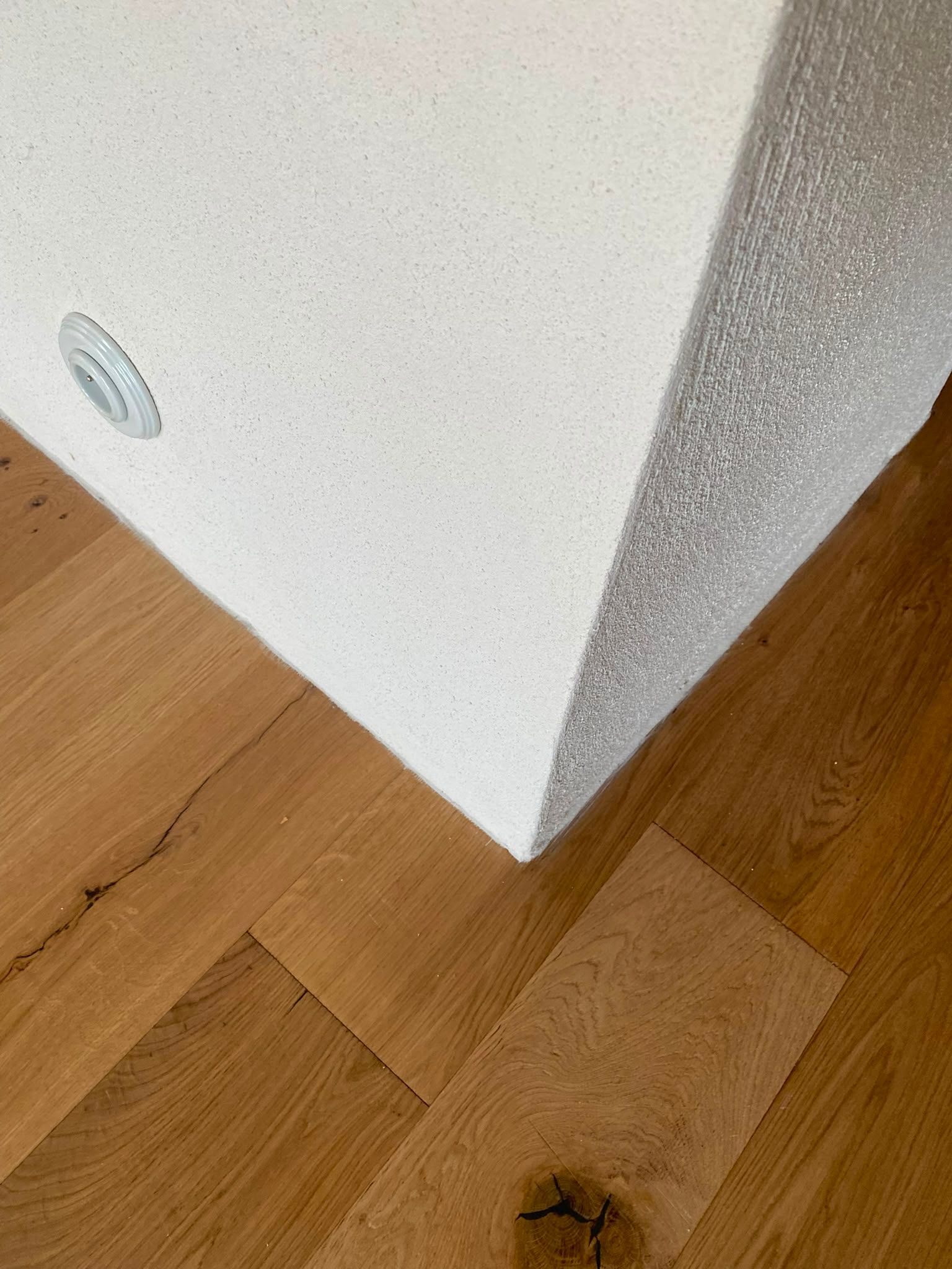 Coin d’un mur blanc au-dessus d’un parquet clair, avec une petite prise près de la plinthe.
