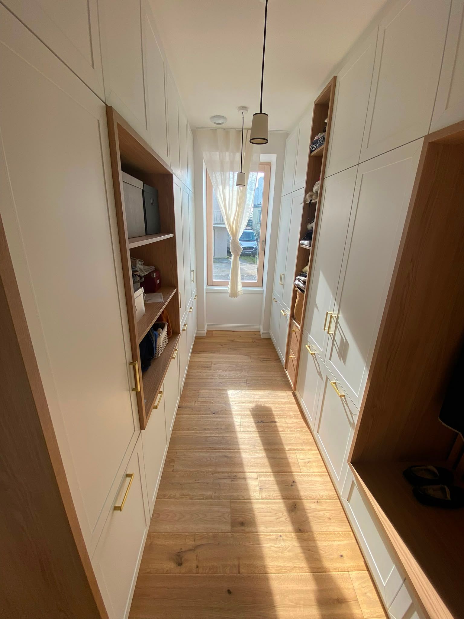 Couloir étroit baigné de soleil, avec un parquet, des murs blancs et des étagères intégrées, menant à une porte lumineuse