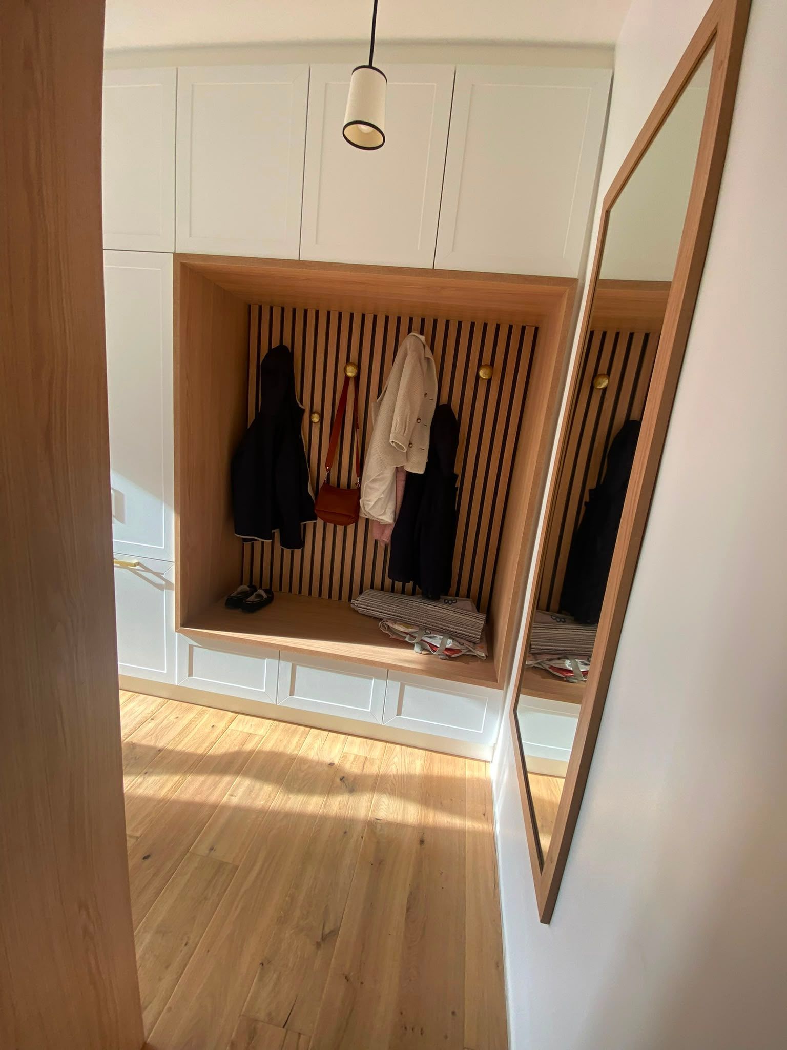 Armoire encastrée en bois avec des vêtements suspendus, un miroir au mur et des rayons de soleil sur le sol.