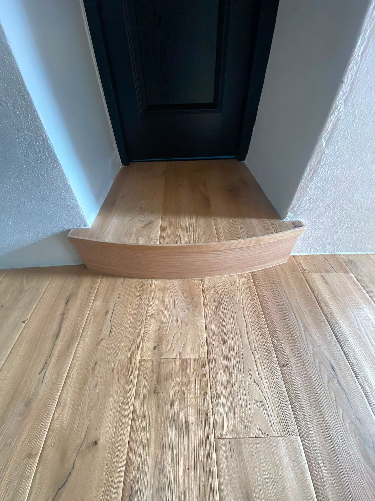 Un plancher en bois mène à une porte sombre, avec une petite marche surélevée au seuil.