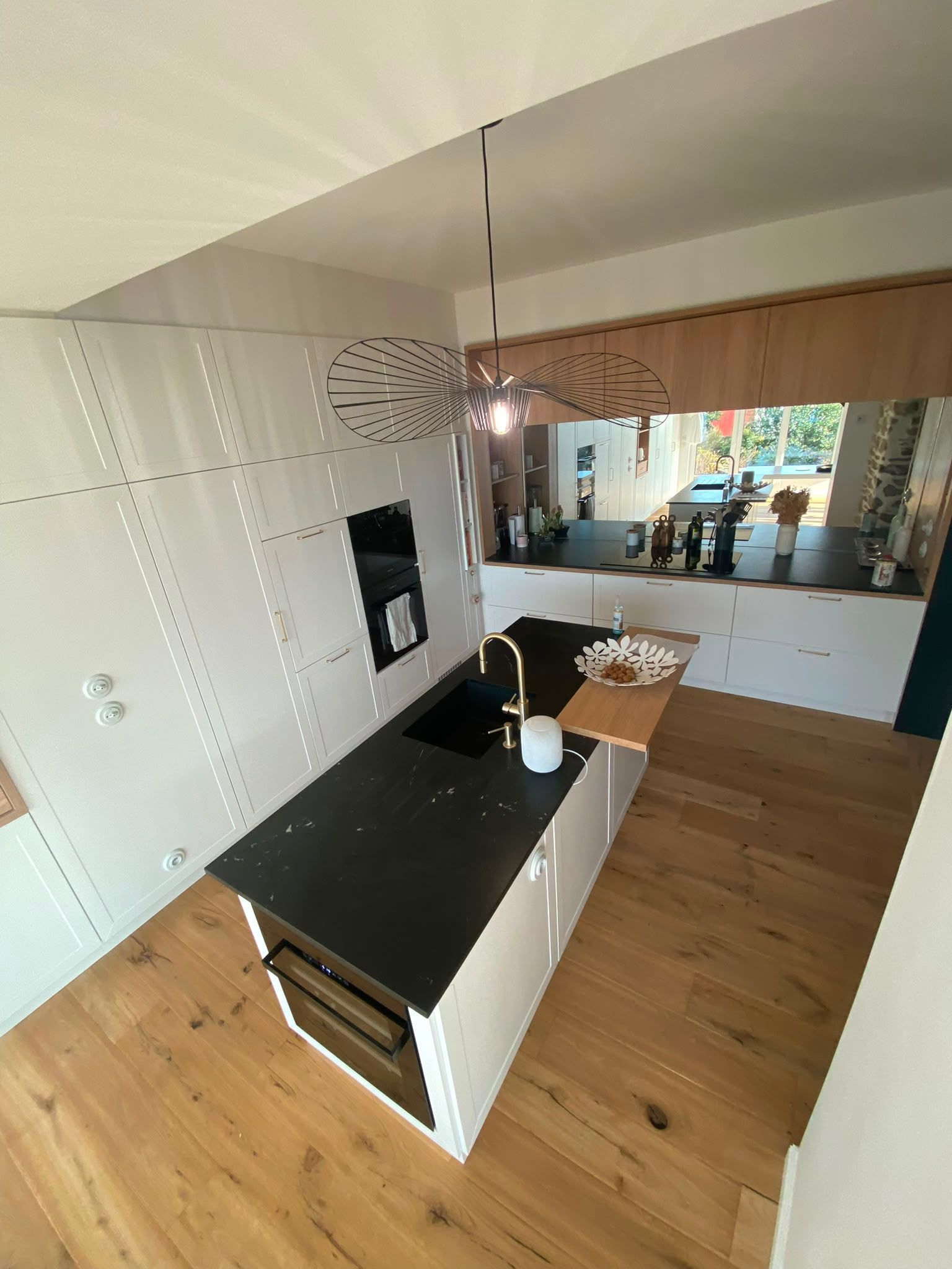 Cuisine moderne avec îlot noir, placards blancs, parquet et salle à manger en arrière-plan