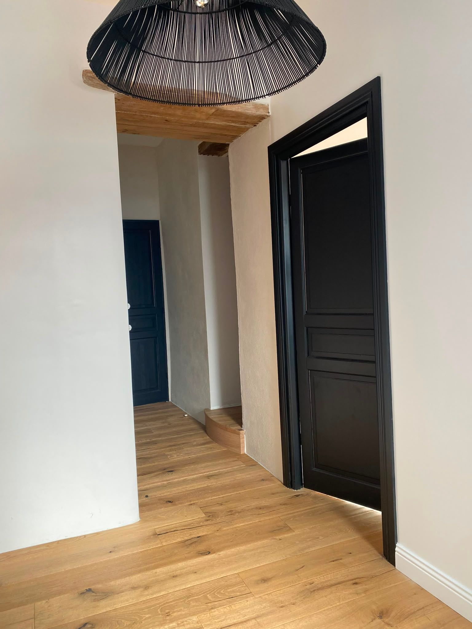 Couloir moderne avec un parquet clair, des murs blancs, des portes noires et une suspension tressée.