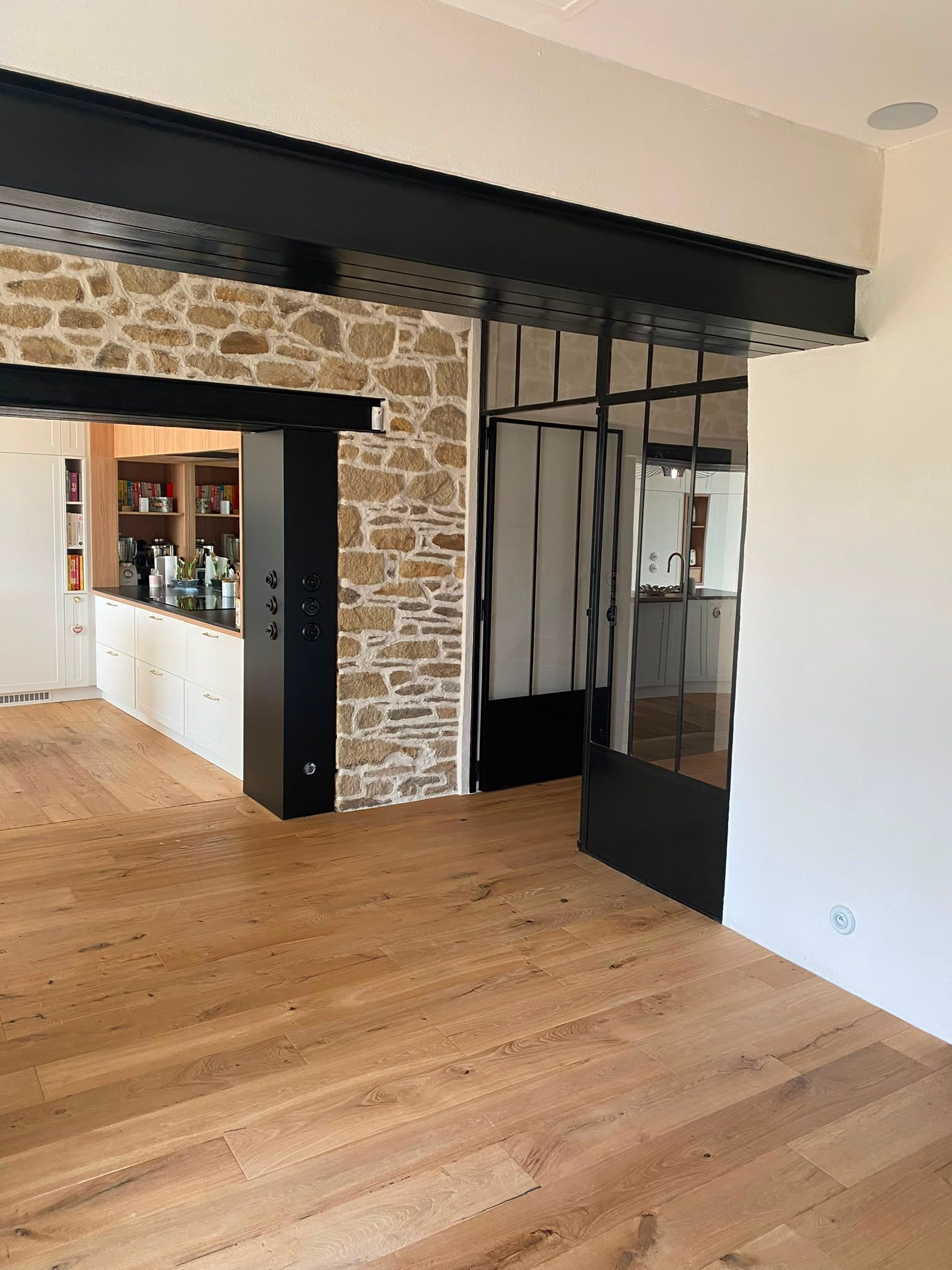 Pièce vide avec parquet clair, embrasure de porte en pierre apparente et cloison vitrée à cadre noir.