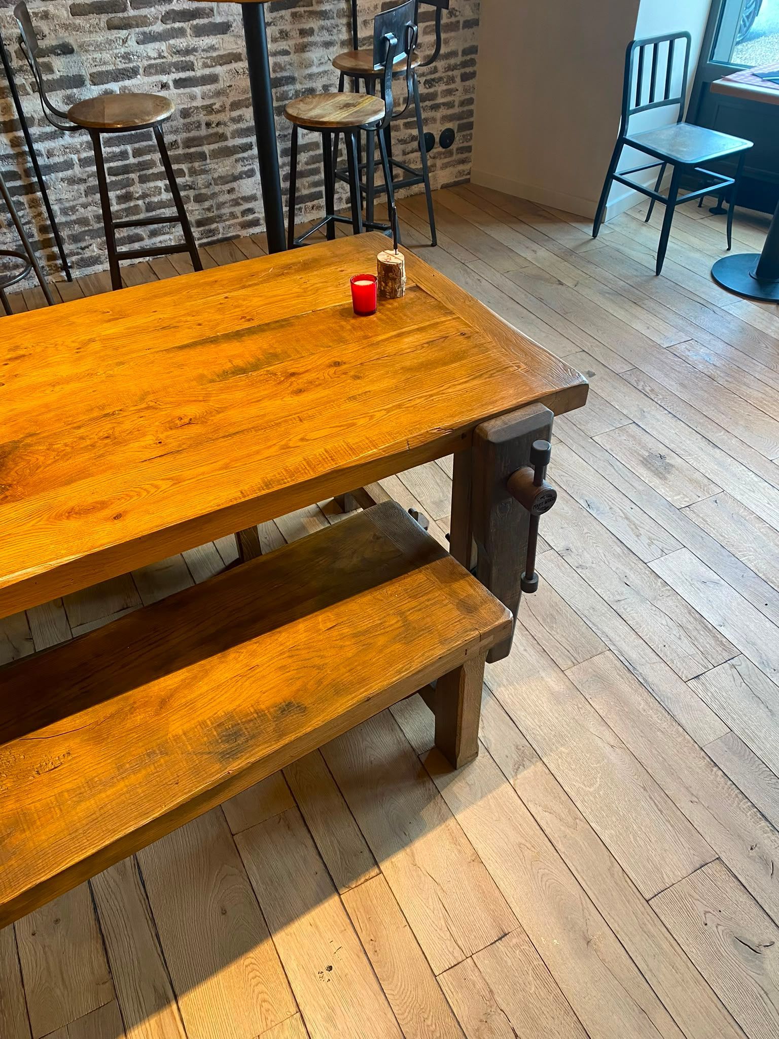 Table et bancs rustiques en bois dans un café lumineux aux murs de briques, avec une petite bougie rouge en centre de table