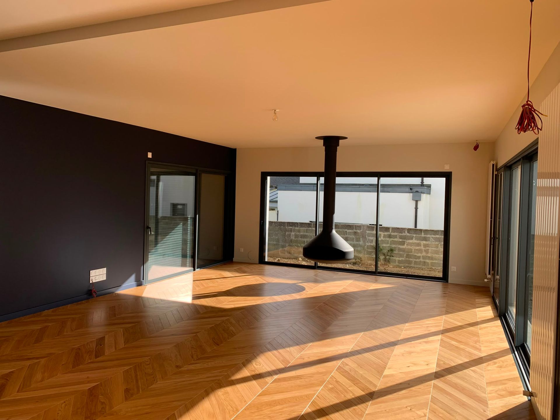 Salon moderne vide avec parquet en bois à chevrons, cheminée noire et grandes portes vitrées ouvrant sur une vue lumineuse