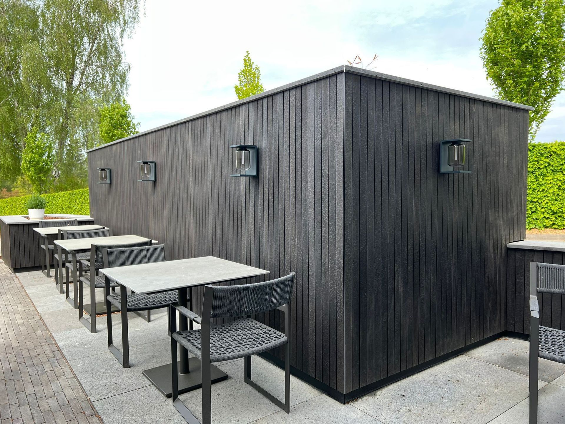 Terrasse extérieure avec espace de détente et tables en bois noir, à côté d’un bâtiment moderne sombre