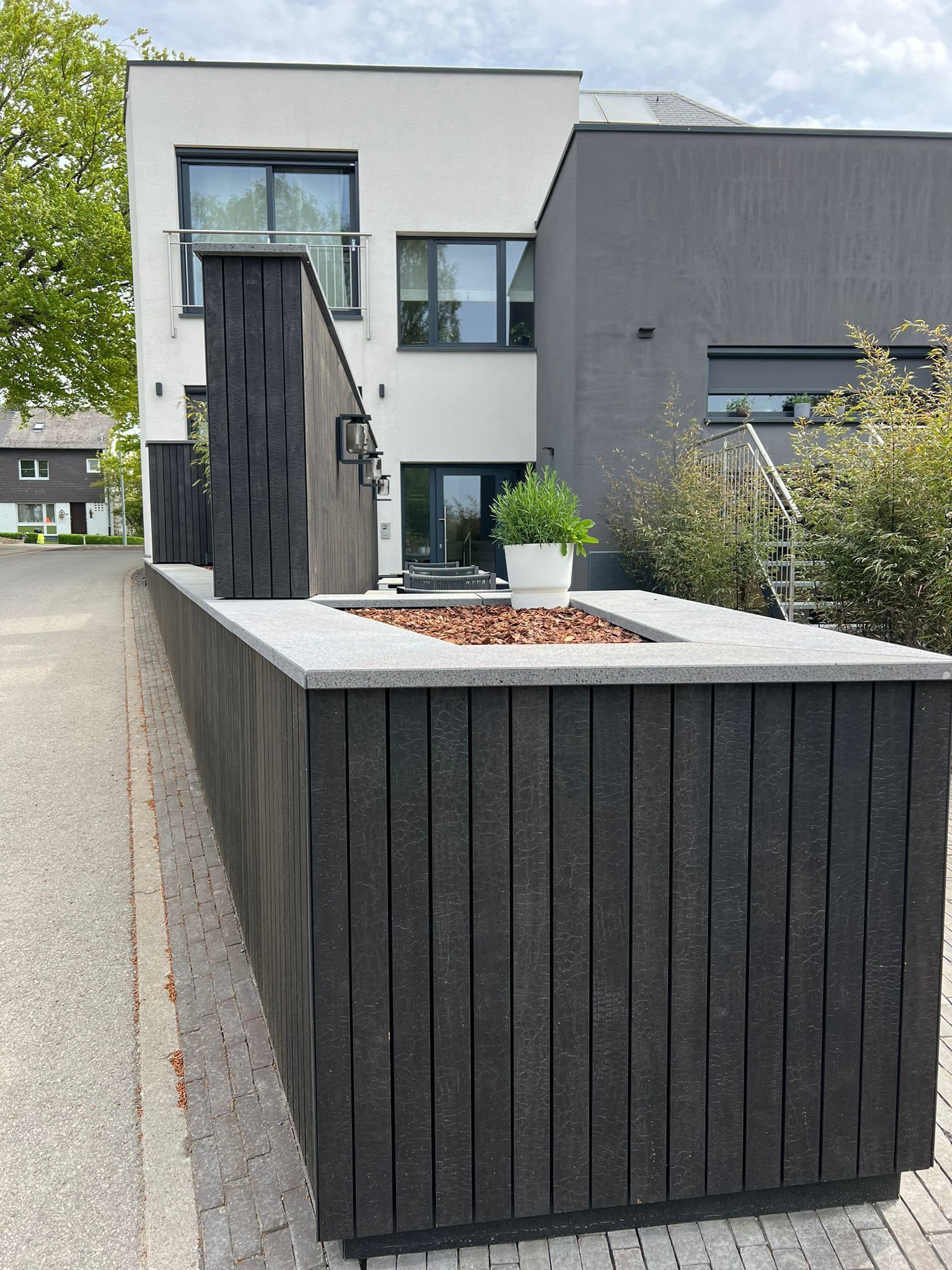 Maison moderne blanche et grise avec une jardinière à lames noires à l’avant et un trottoir au premier plan