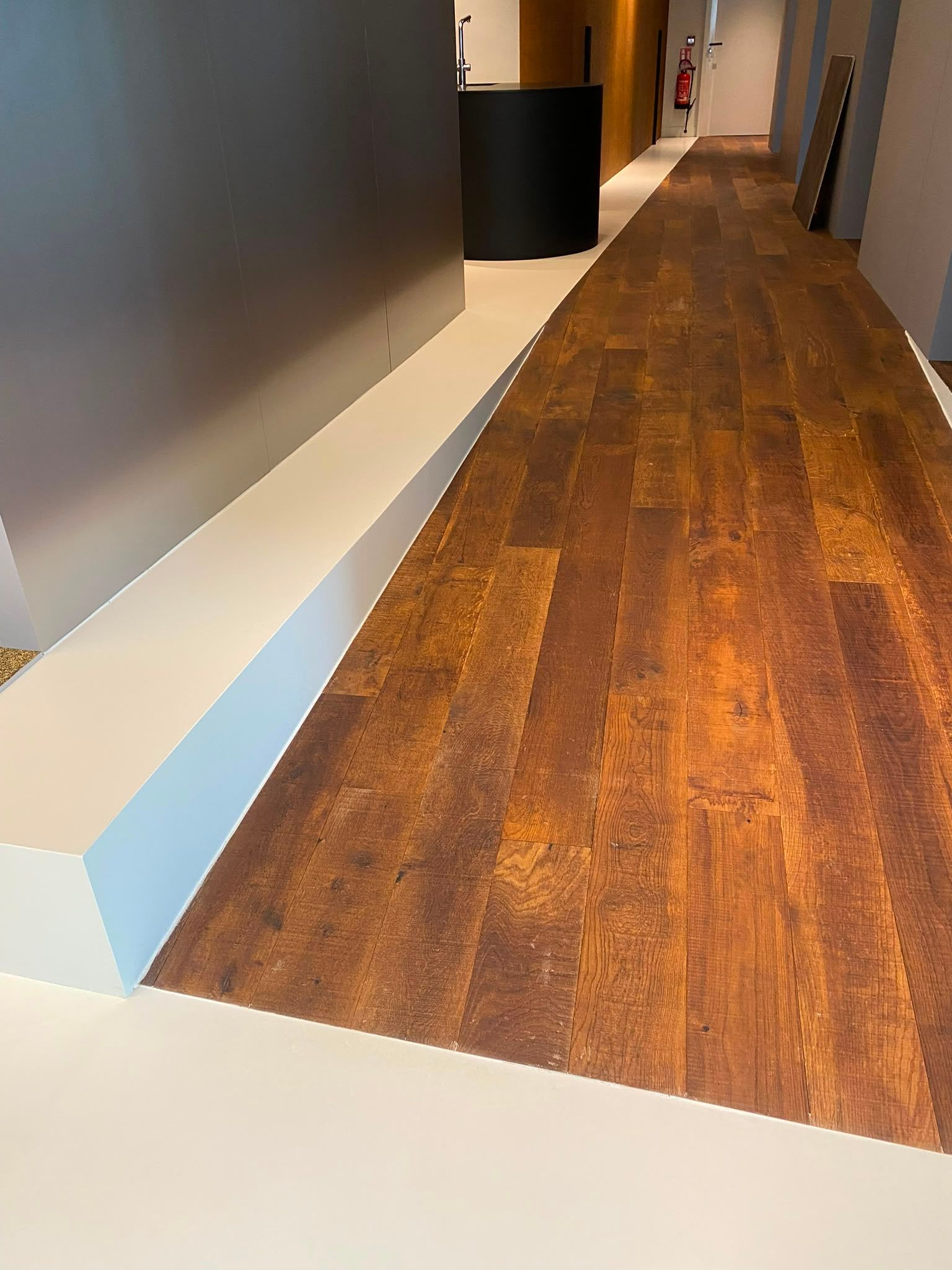 Couloir vide avec un parquet en bois, des murs gris et une banquette blanche le long du mur gauche.