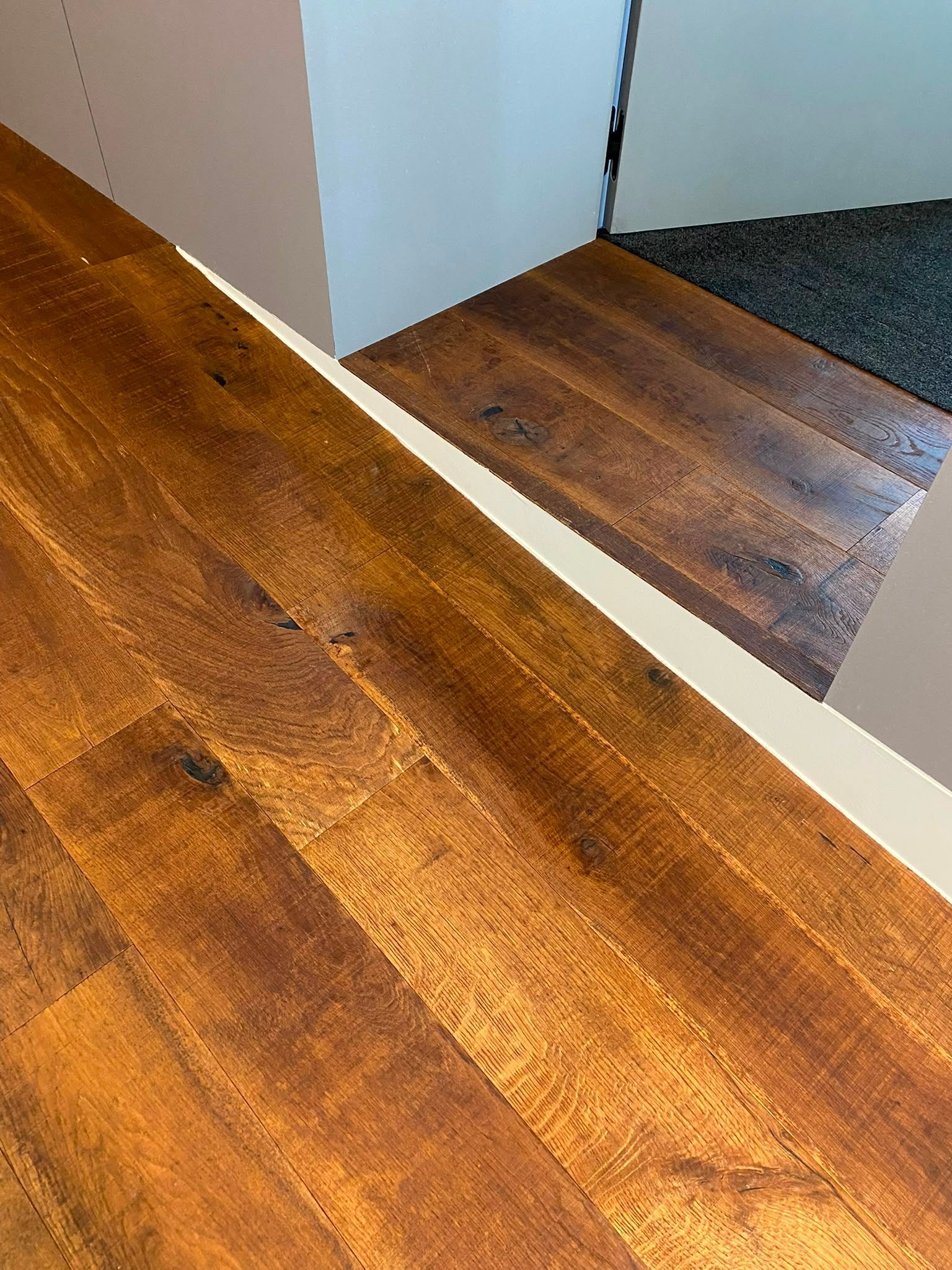 Parquet en bois au pied d’un mur miroir, reflétant un couloir et une zone recouverte de moquette sombre.