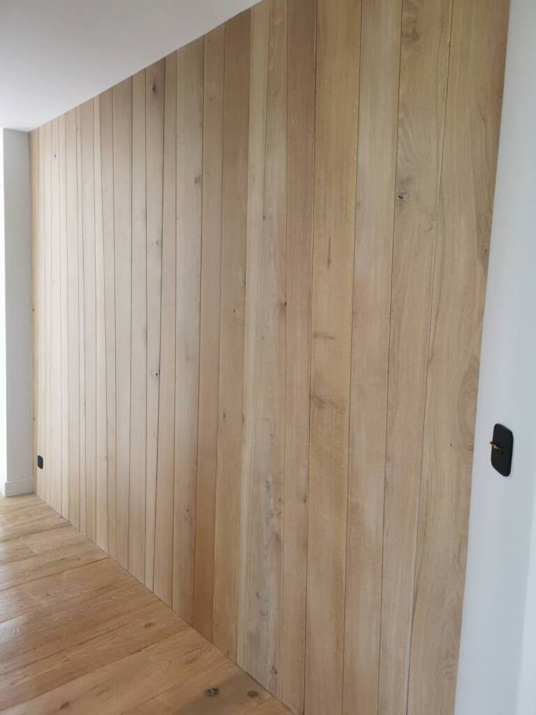 Couloir moderne avec un revêtement mural et un parquet en bois clair, à côté d’un mur blanc et d’une porte.