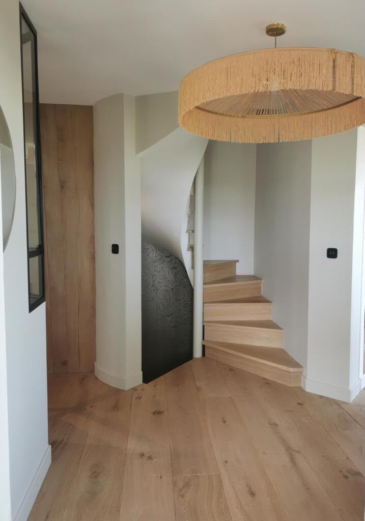 Couloir moderne avec parquet clair, suspension tressée et escalier arrondi près d’une porte en bois.