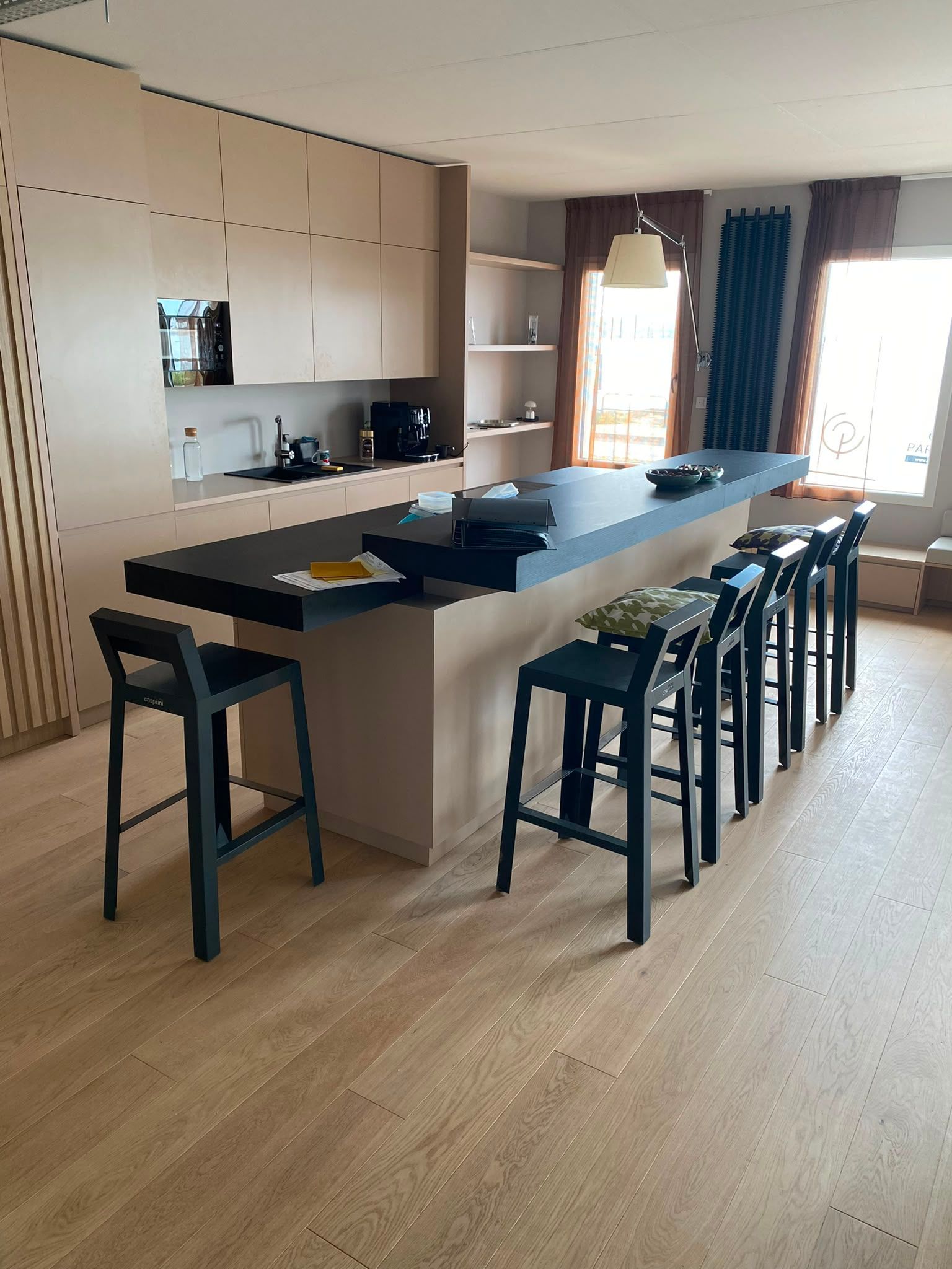 Cuisine moderne avec un grand îlot, des tabourets noirs, des placards en bois clair et une fenêtre avec des rideaux sombres