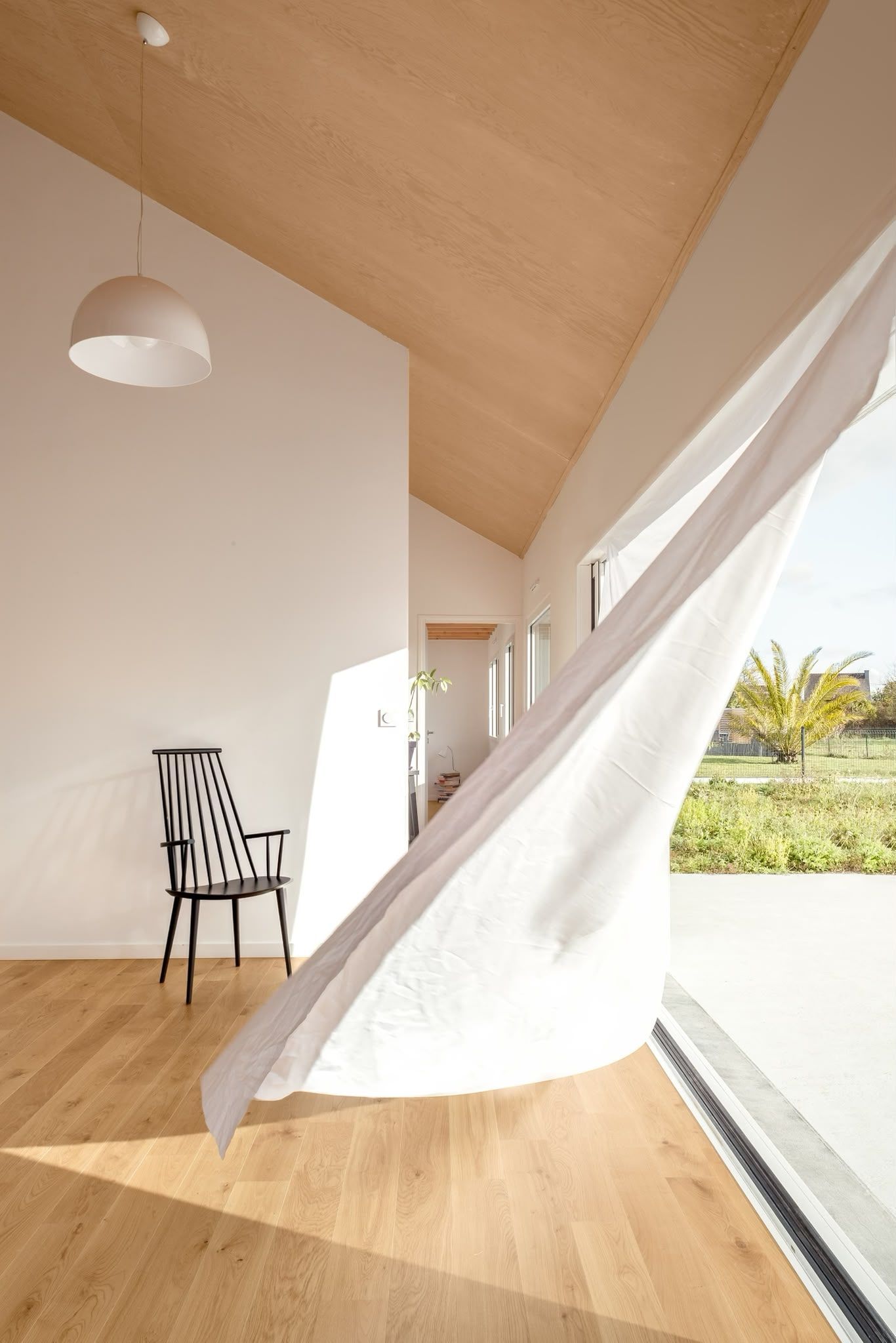 Patio moderne baigné de soleil, avec des rideaux blancs, un plafond en bois, une chaise et une vue sur la verdure extérieure