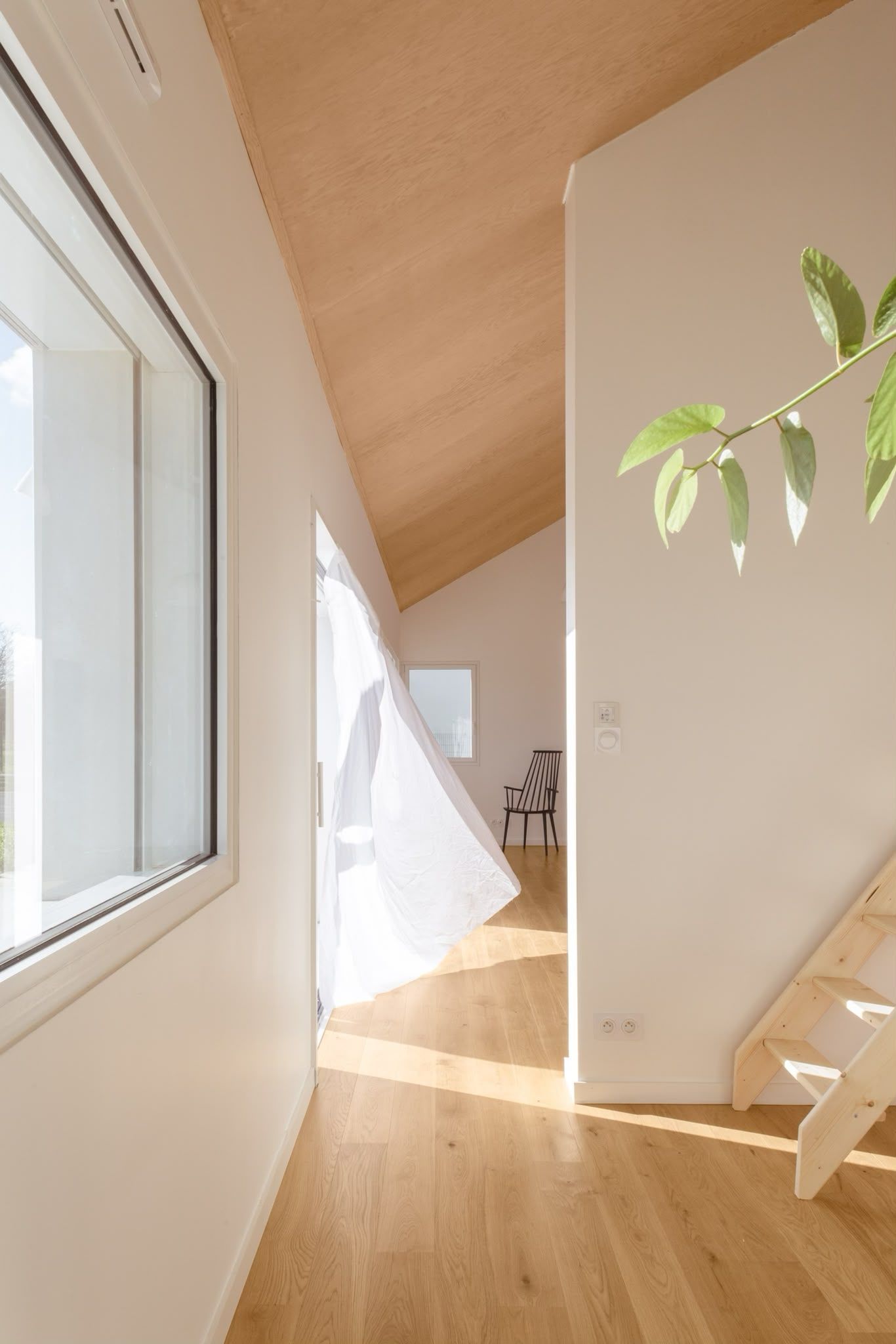 Couloir lumineux avec parquet, grande fenêtre, plafond en pente et rideau transparent baigné de soleil.