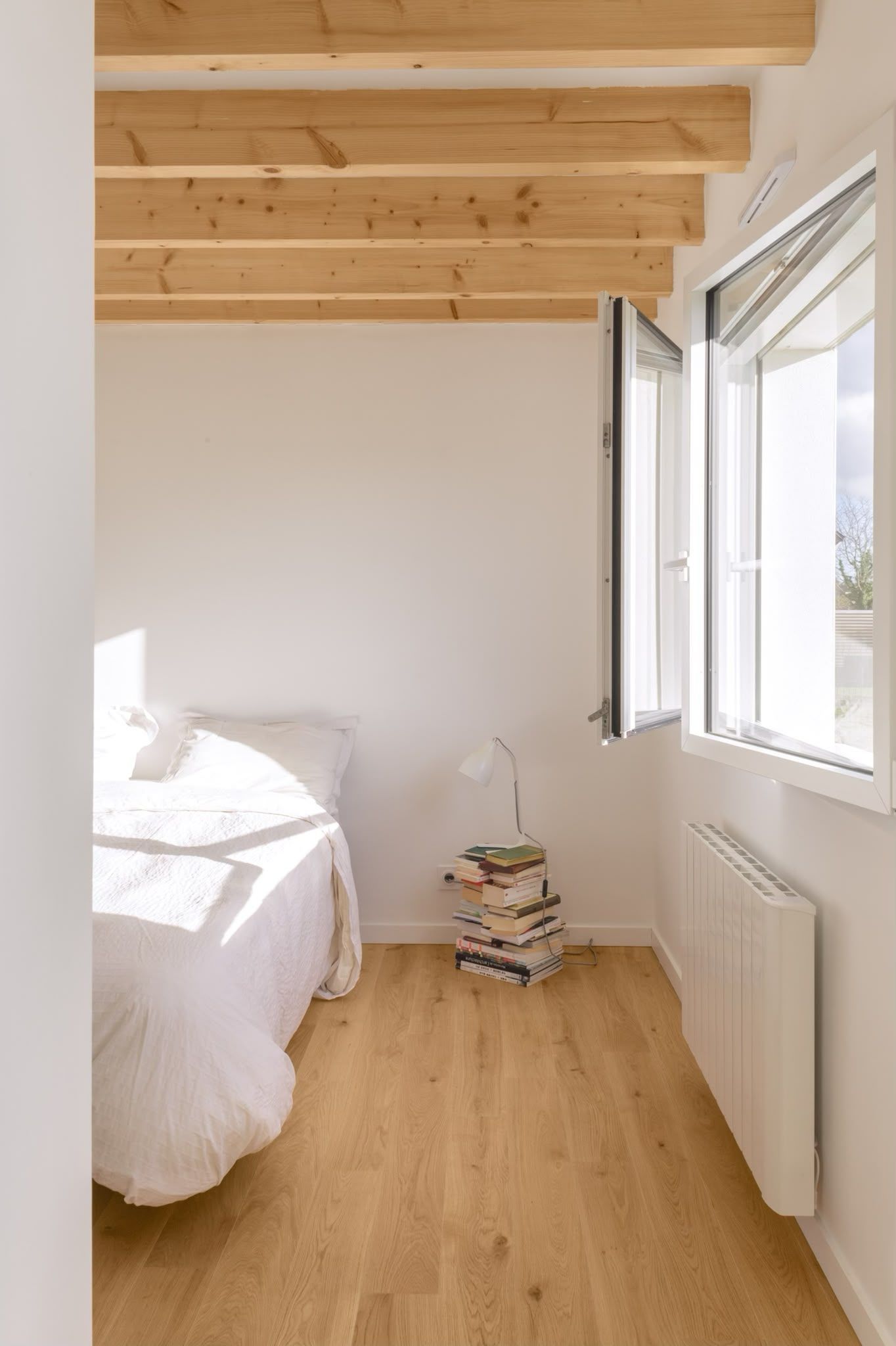 Chambre lumineuse et minimaliste avec un lit blanc, un plafond en bois, une fenêtre ouverte et des livres empilés sur le sol