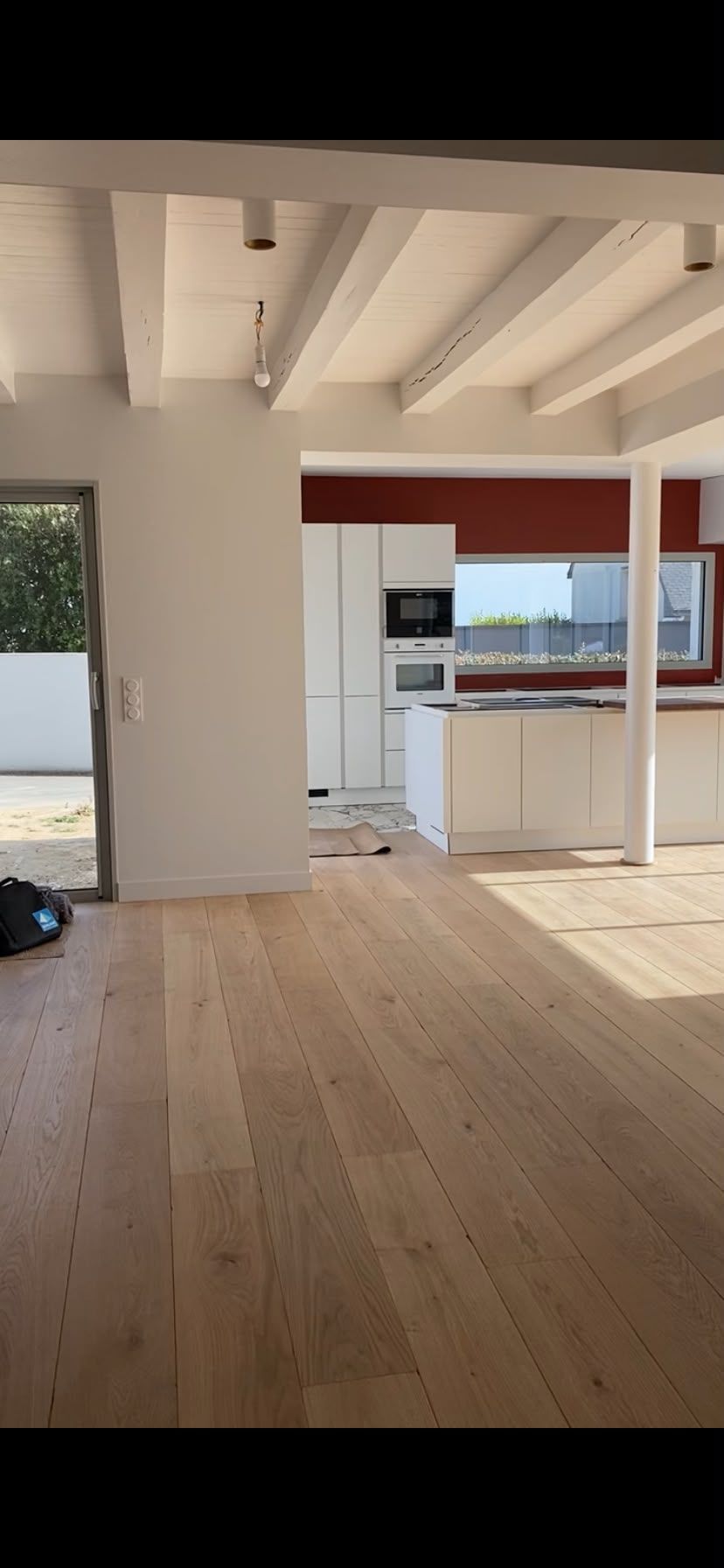 Pièce moderne vide avec parquet clair, poutres blanches et cuisine donnant sur un extérieur lumineux