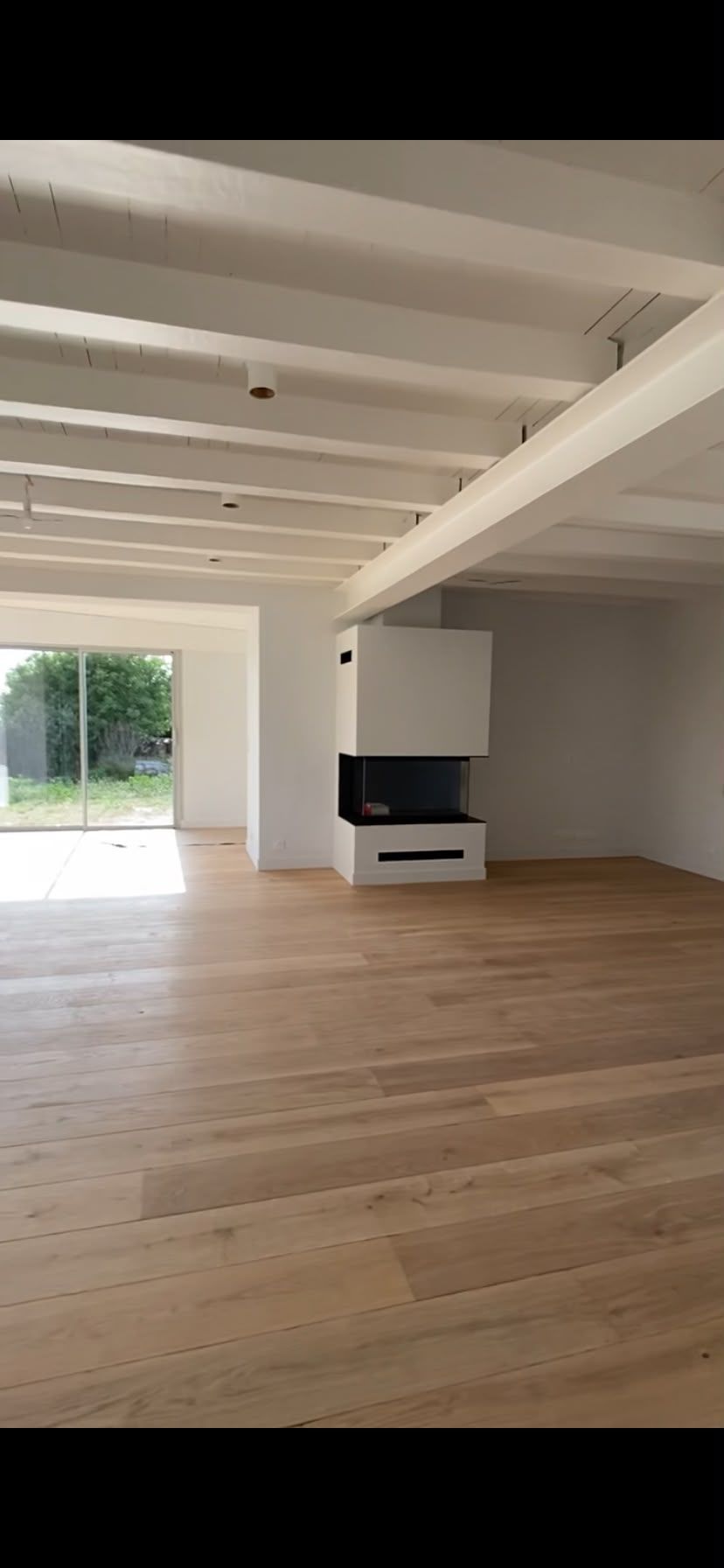 Salon vide et lumineux, avec parquet, poutres apparentes et vue sur l’extérieur depuis la fenêtre