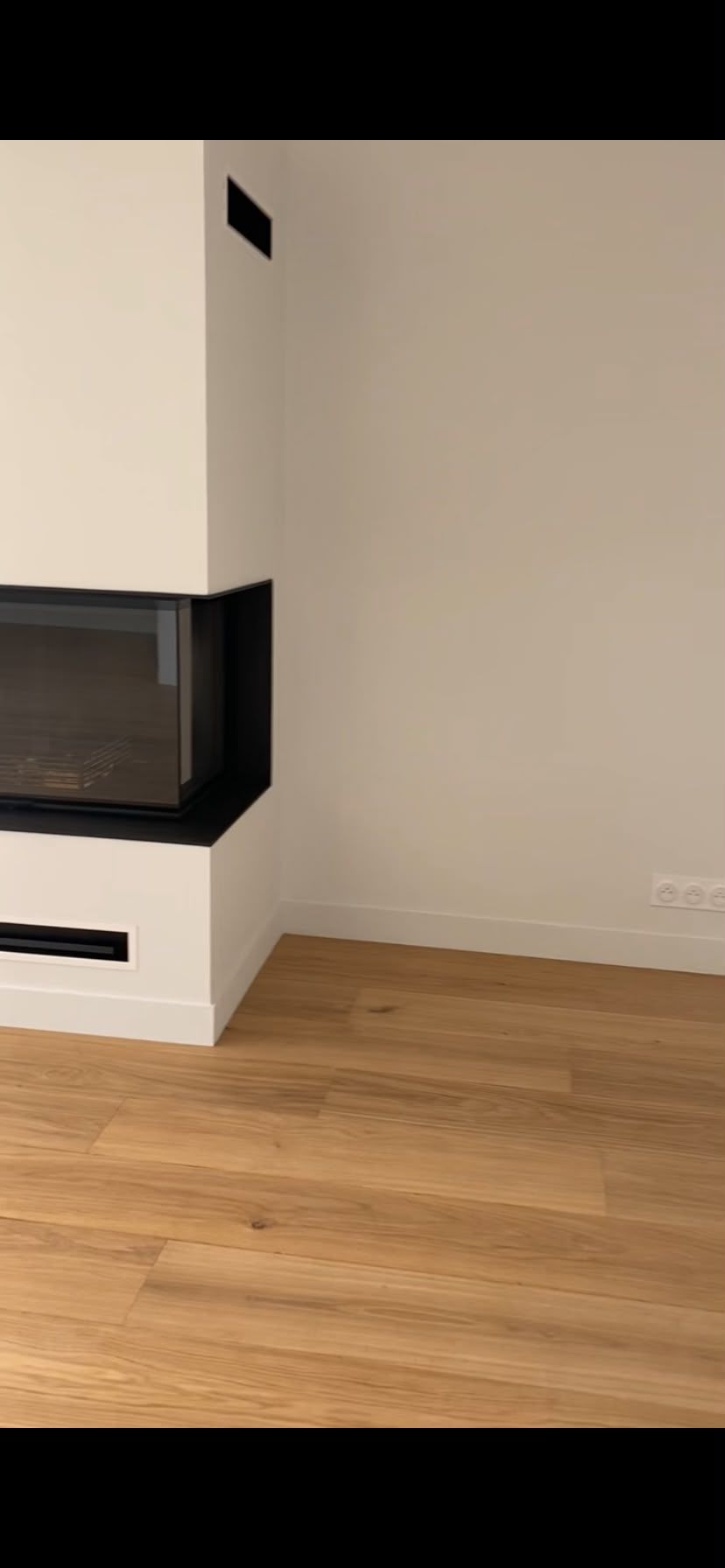 Coin d’une pièce moderne vide, avec des murs blancs, un parquet en bois et une cheminée noire.