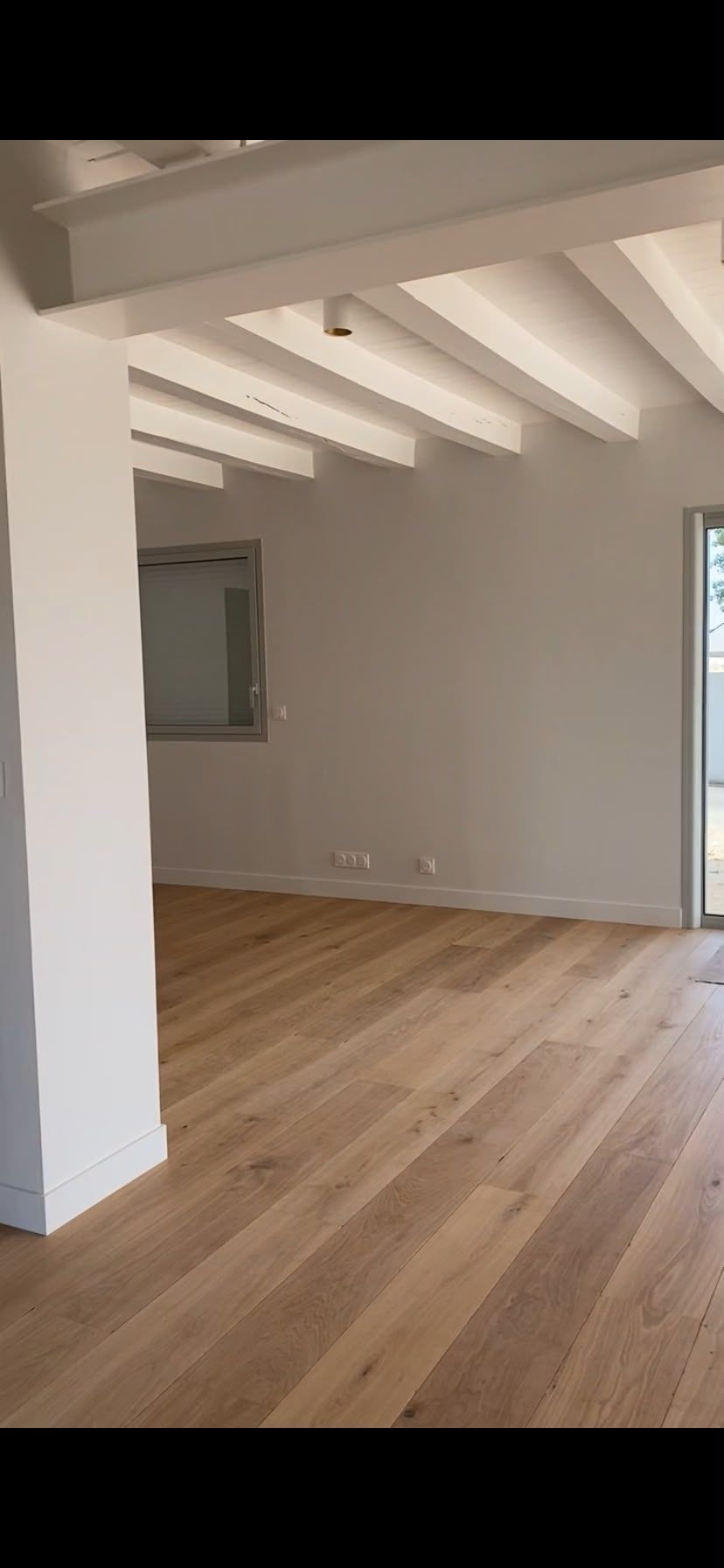Pièce moderne vide avec un parquet clair, des murs blancs, des poutres apparentes et une fenêtre.