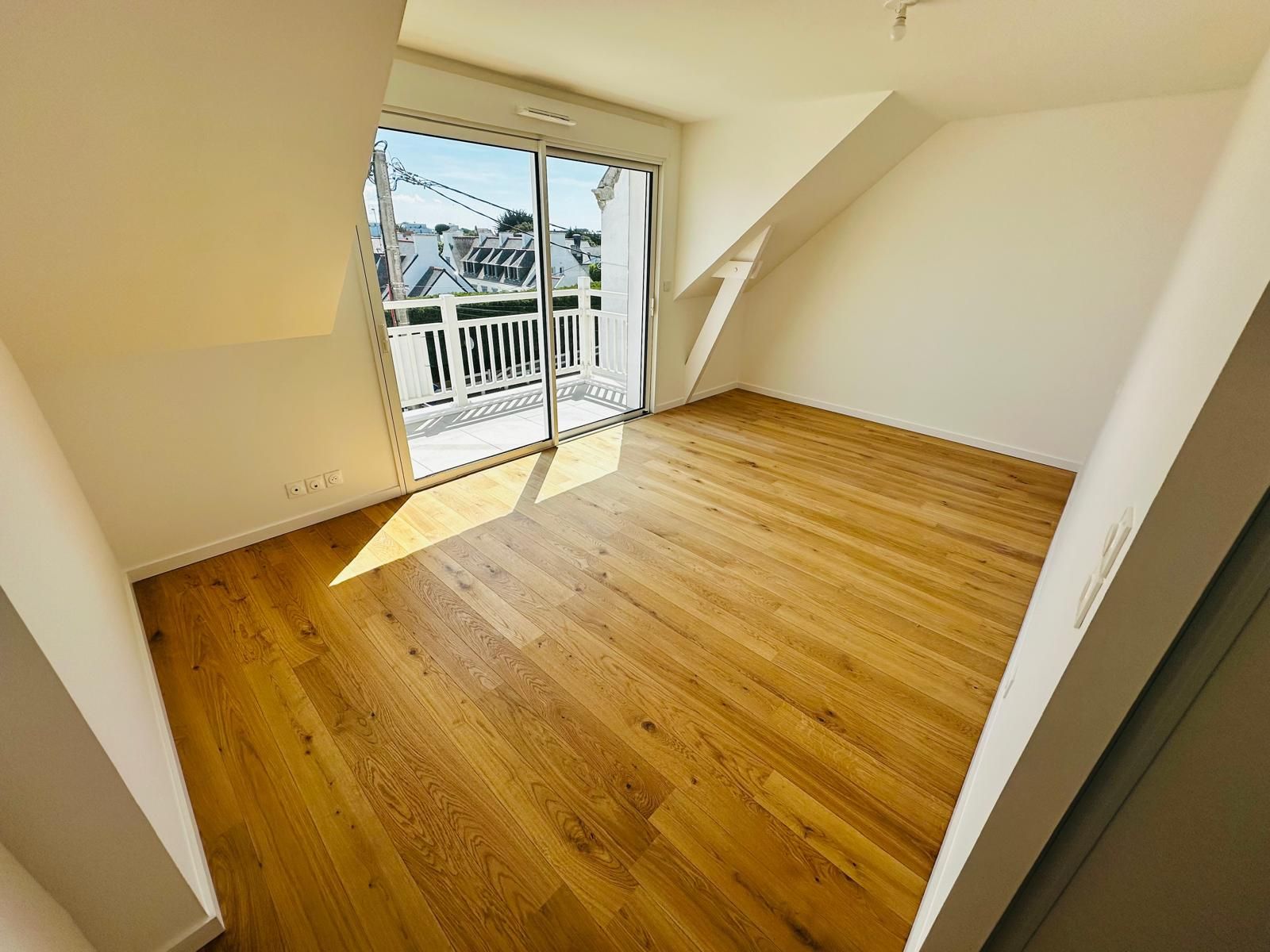 Pièce vide baignée de soleil, avec un parquet, un plafond en pente et une porte vitrée ouverte sur un balcon.