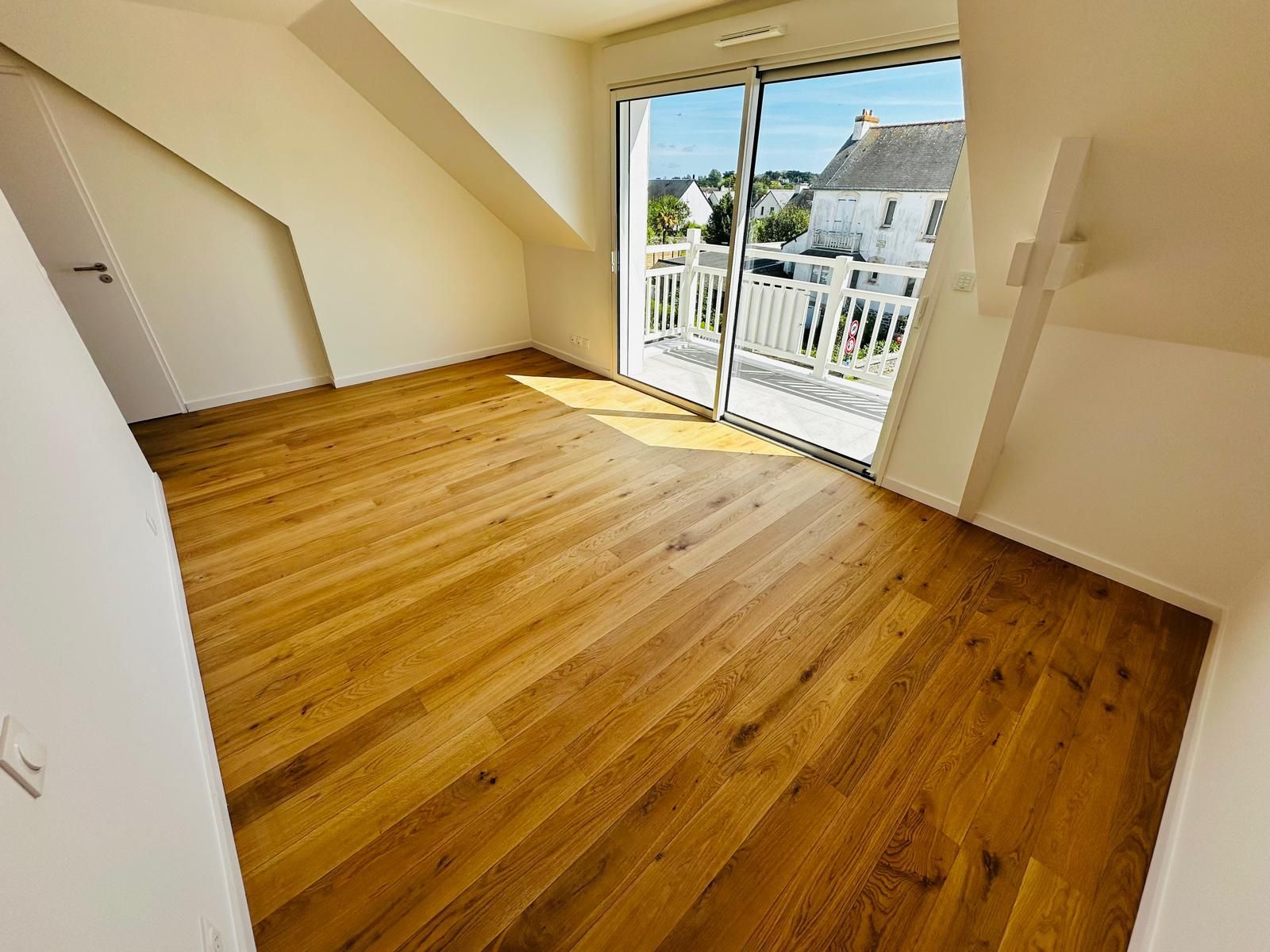 Pièce mansardée vide avec parquet et portes coulissantes vitrées donnant sur un balcon, baignée de soleil
