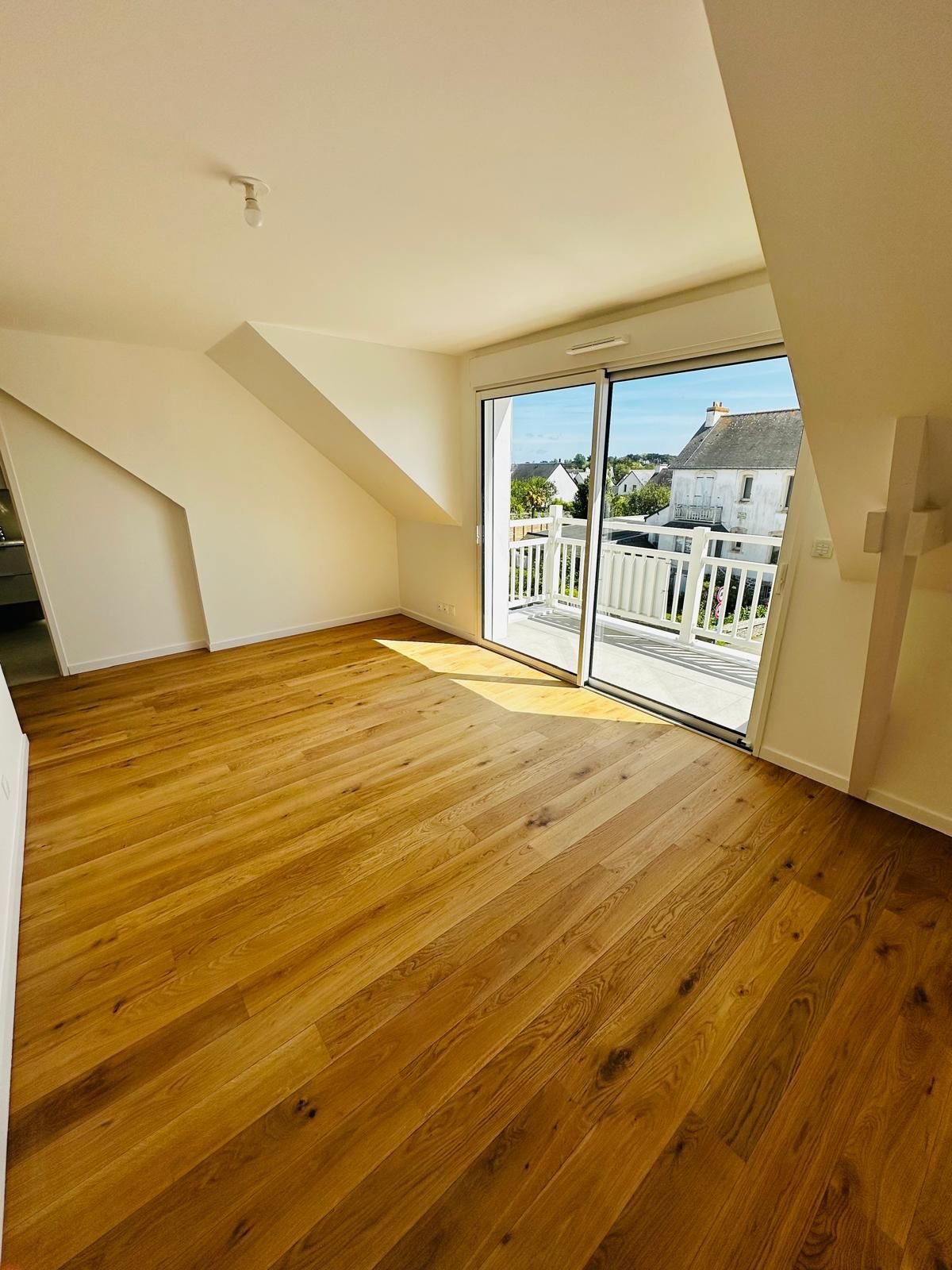 Pièce lumineuse et vide avec un parquet, un plafond mansardé et de grandes baies vitrées coulissantes donnant sur un balcon.