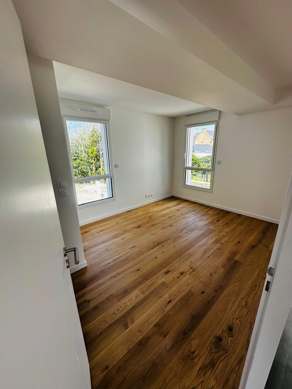 Pièce blanche vide avec plafond mansardé, parquet et deux fenêtres donnant sur la verdure extérieure