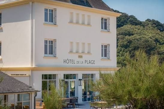 Bâtiment blanc d’un hôtel avec des volets bleus et une enseigne indiquant « Hôtel de la Plage », entouré de verdure.