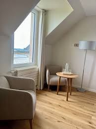 Chambre mansardée cosy avec un fauteuil, une table d’appoint, un lampadaire et une grande fenêtre donnant sur l’eau