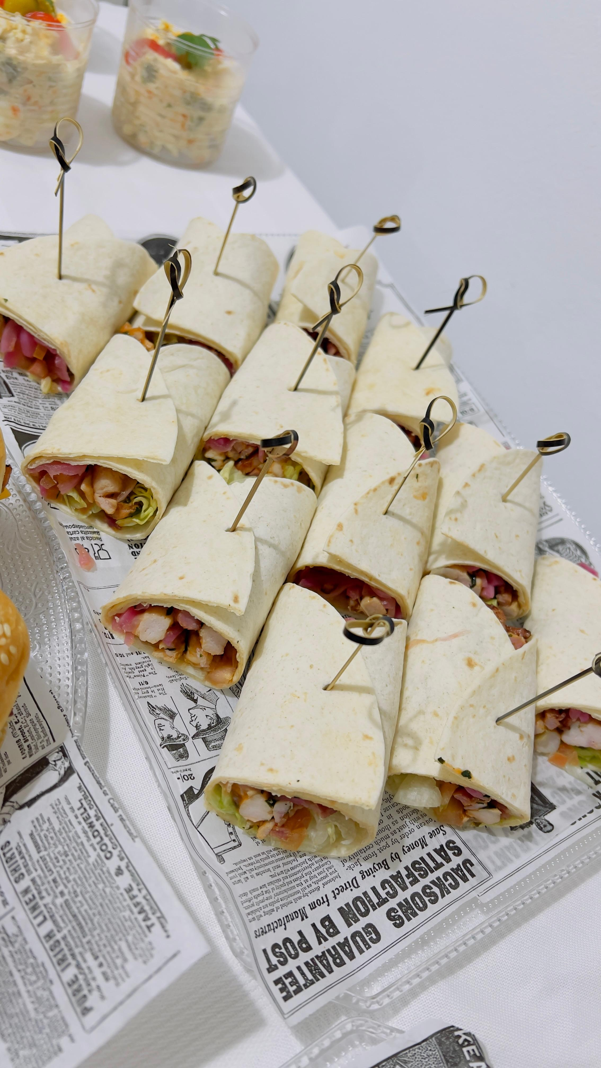 Variedad de sándwiches enrollados sobre una mesa, sujetos con palillos de dientes y dispuestos en bandejas forradas.