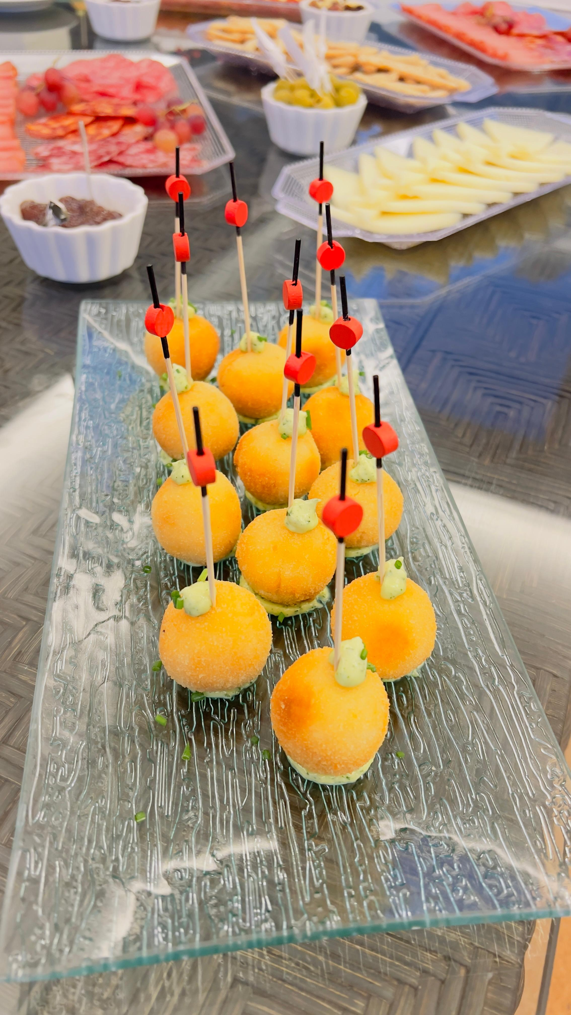 Bocados de fruta naranja ensartados en brochetas sobre una bandeja de cristal, con bandejas de buffet al fondo.