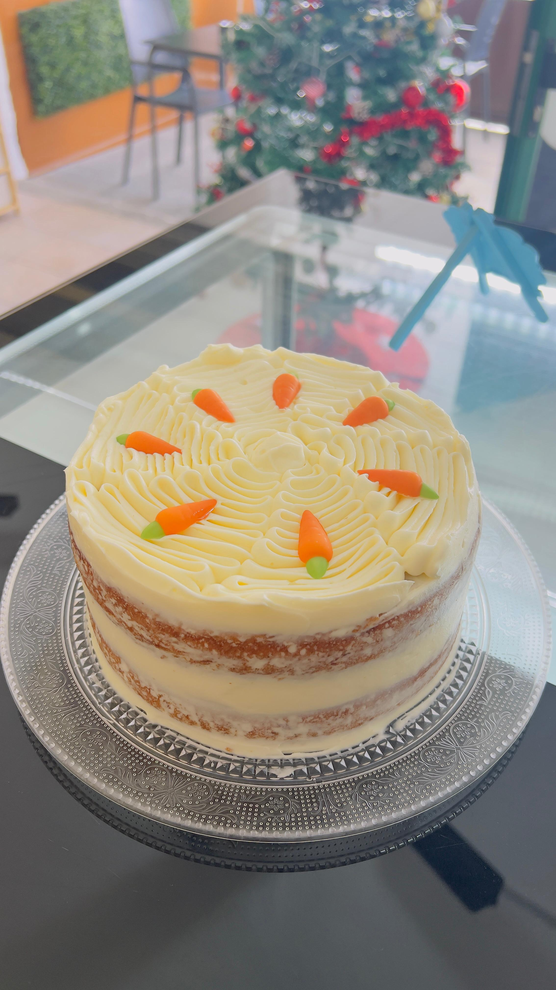 Pastel redondo de crema en capas con guarnición de naranja sobre una mesa de cristal, frente a una ventana con plantas.