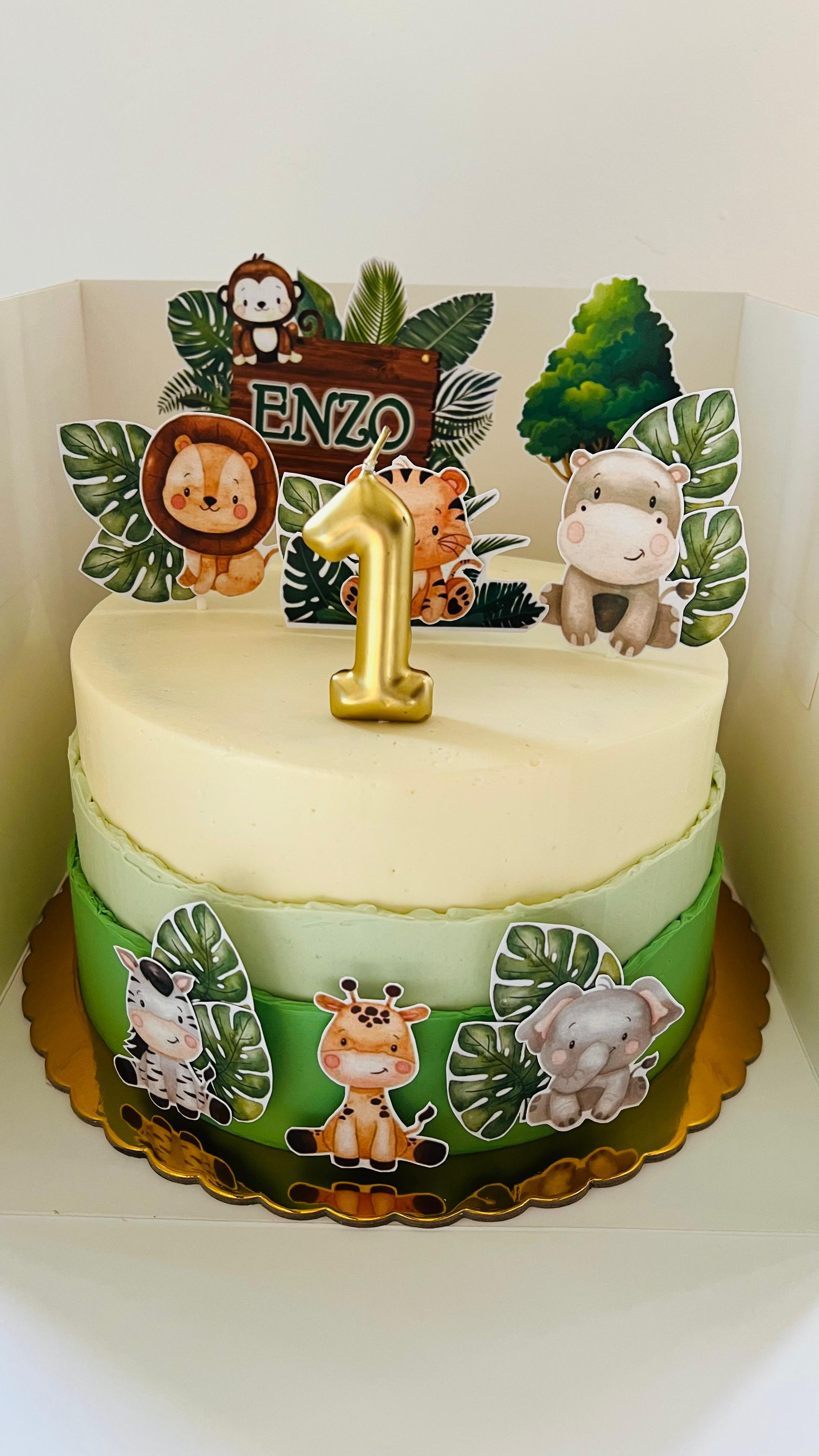 Tarta blanca de primer cumpleaños con adornos de animales de la selva, el número 1 dorado y decoraciones de hojas verdes.