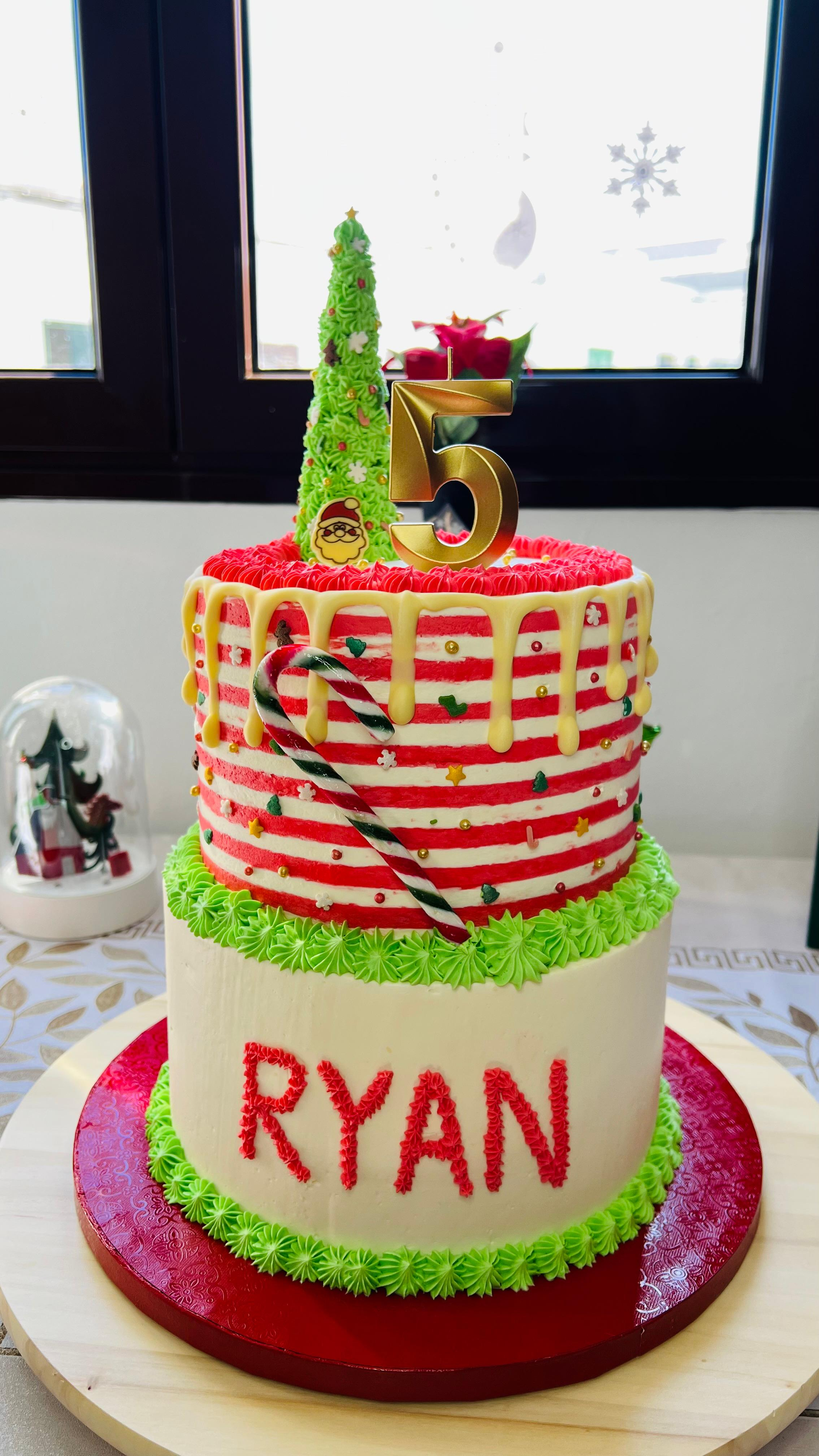 Tarta de cumpleaños de dos pisos con glaseado rojo, verde y blanco, el nombre Ryan y un adorno dorado con el número 5.
