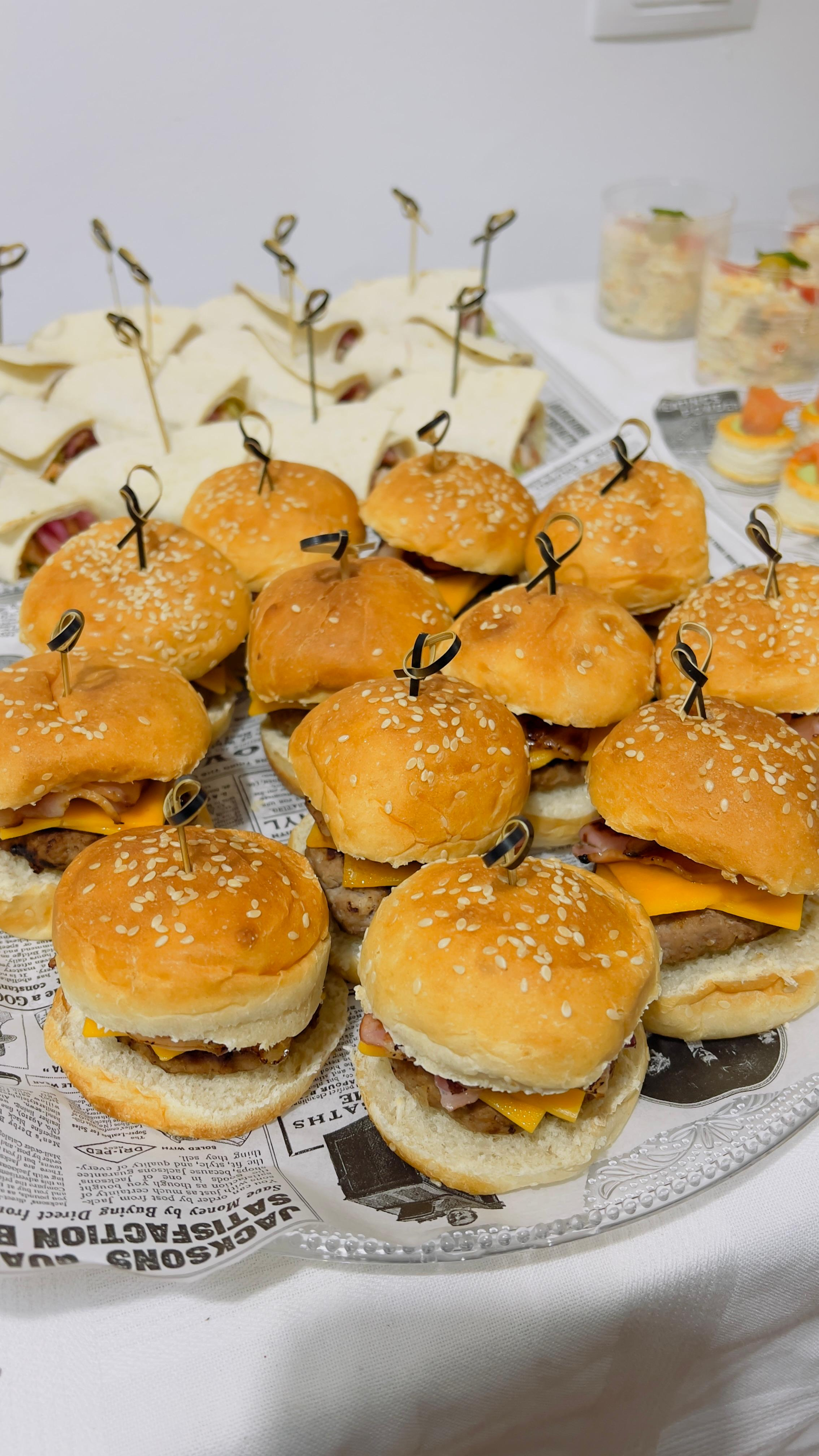 Bandeja de mini hamburguesas con semillas de sésamo sobre una mesa, con adornos de palillos de dientes al fondo.